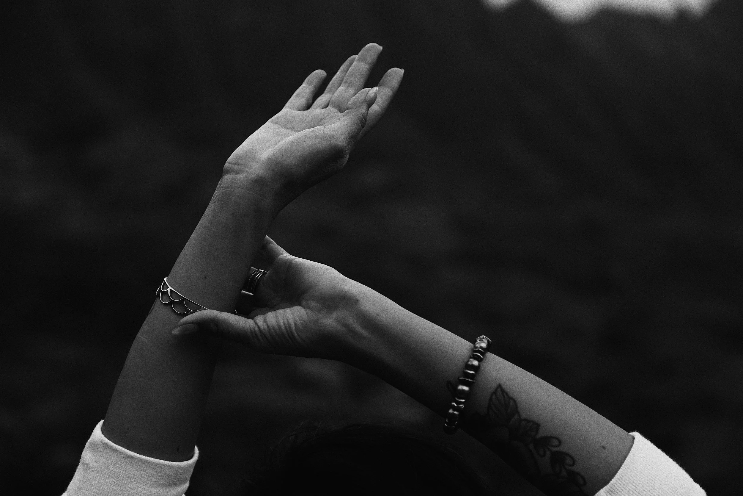 Black and white photograph of two hands gently touching, with one hand raised and the other supporting it. The person with the raised hand has a tattoo of a flower on their forearm, is wearing rings and a bracelet, and has a long-sleeved shirt. The b