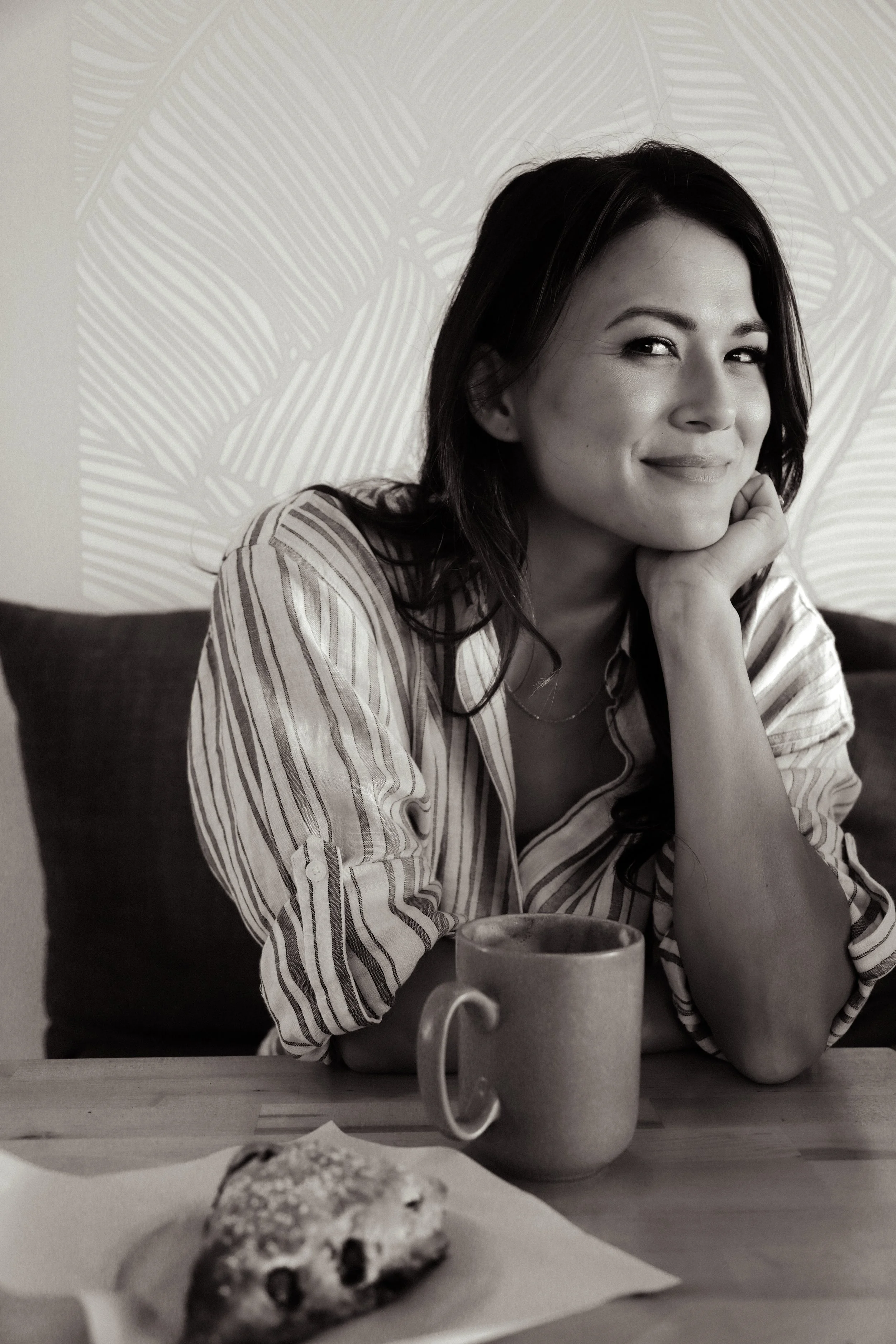 A woman with dark hair sitting at a table with a coffee mug and a pastry, smiling and resting her head on her hand.