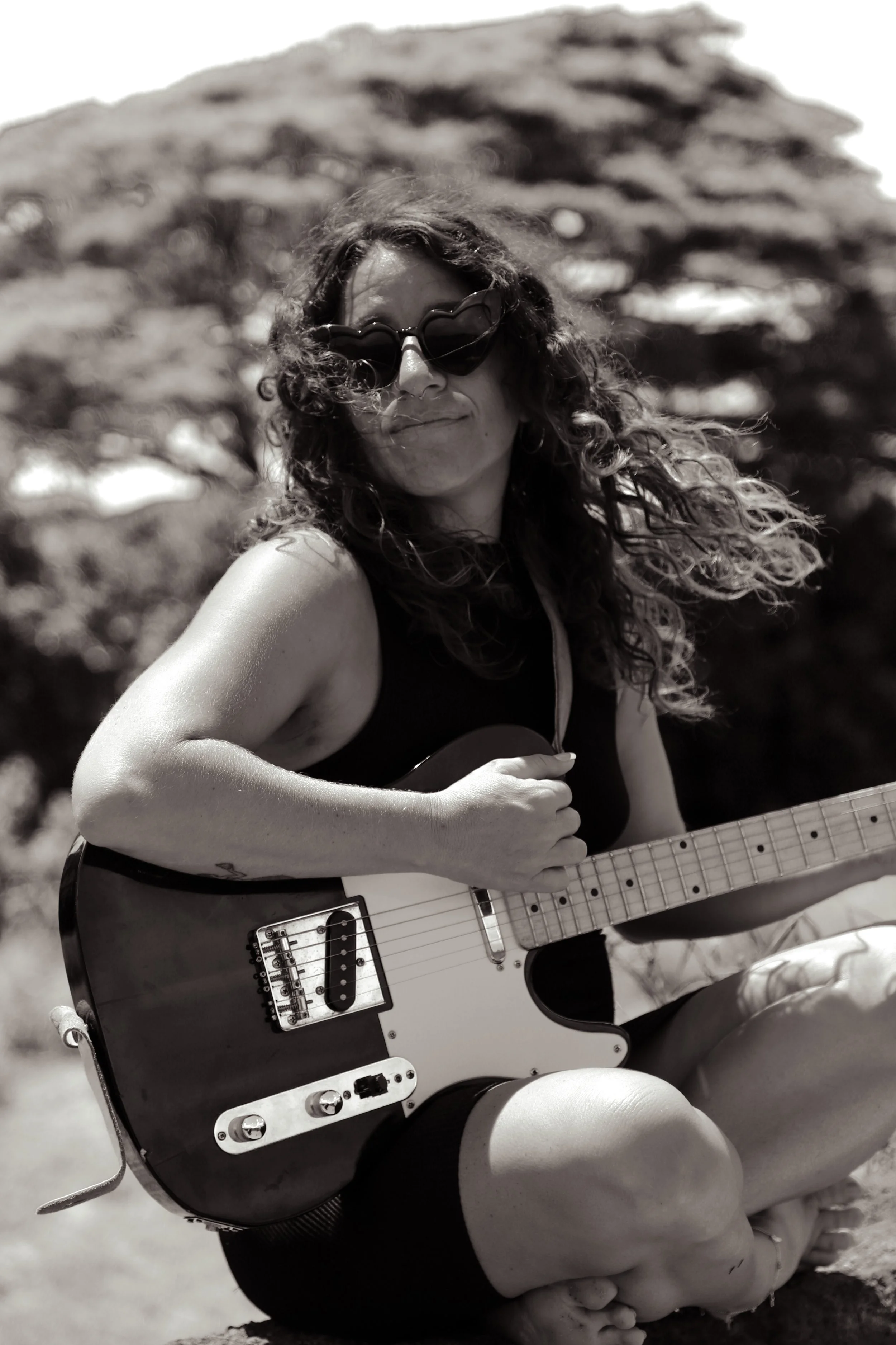 A woman with curly hair wearing sunglasses and a sleeveless top, sitting outdoors, playing an electric guitar with a black and white finish.