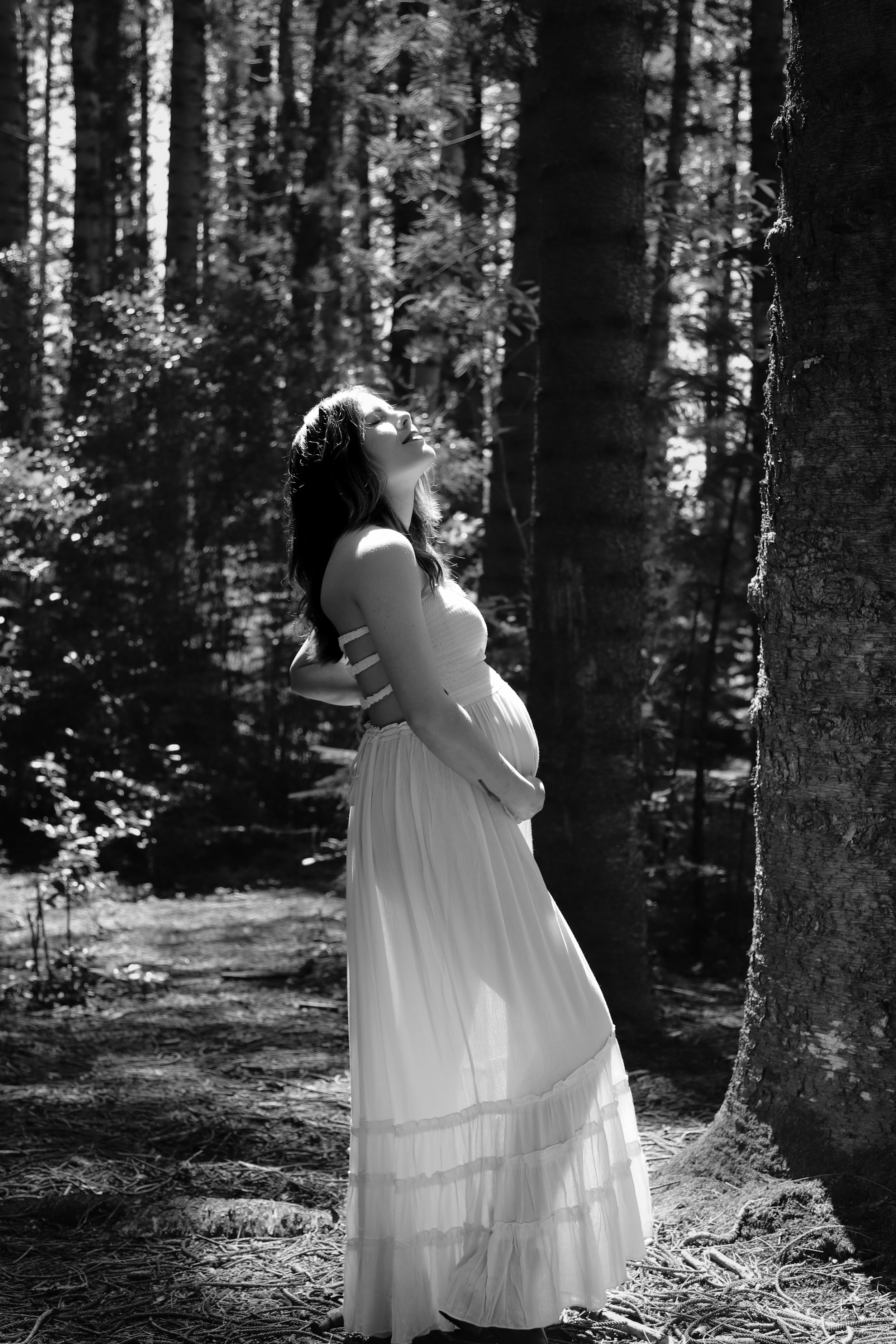 A pregnant woman in a white dress standing in a forest, with sunlight filtering through trees, and her face raised towards the sky.