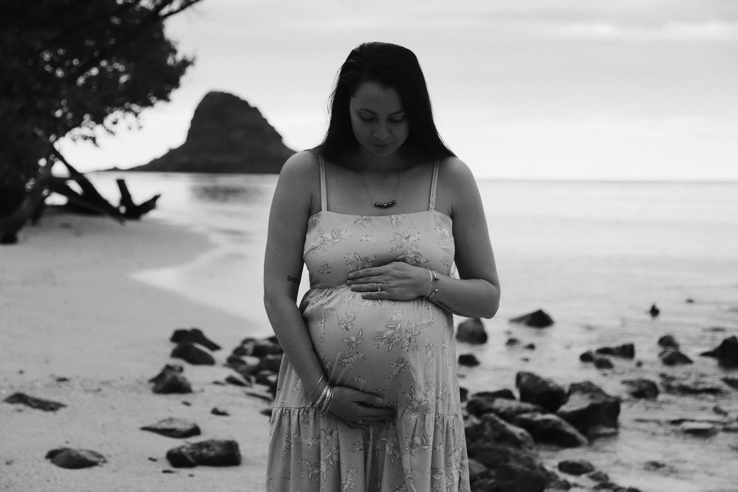 A pregnant woman in a floral dress standing on a beach with rocks, holding her belly, with a tropical island and trees in the background, in black and white.