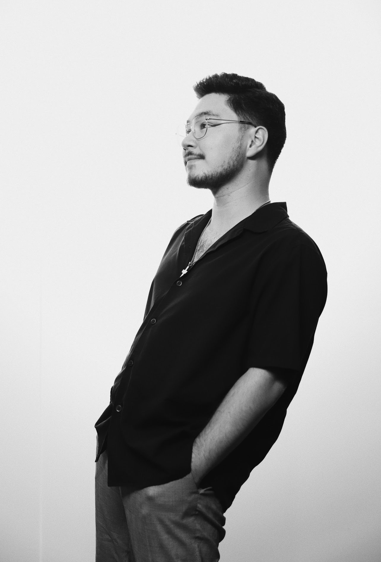 Black and white photo of a man with glasses, beard, dressed in a dark shirt with his hands in his pockets, looking to the side against a plain light-colored background.