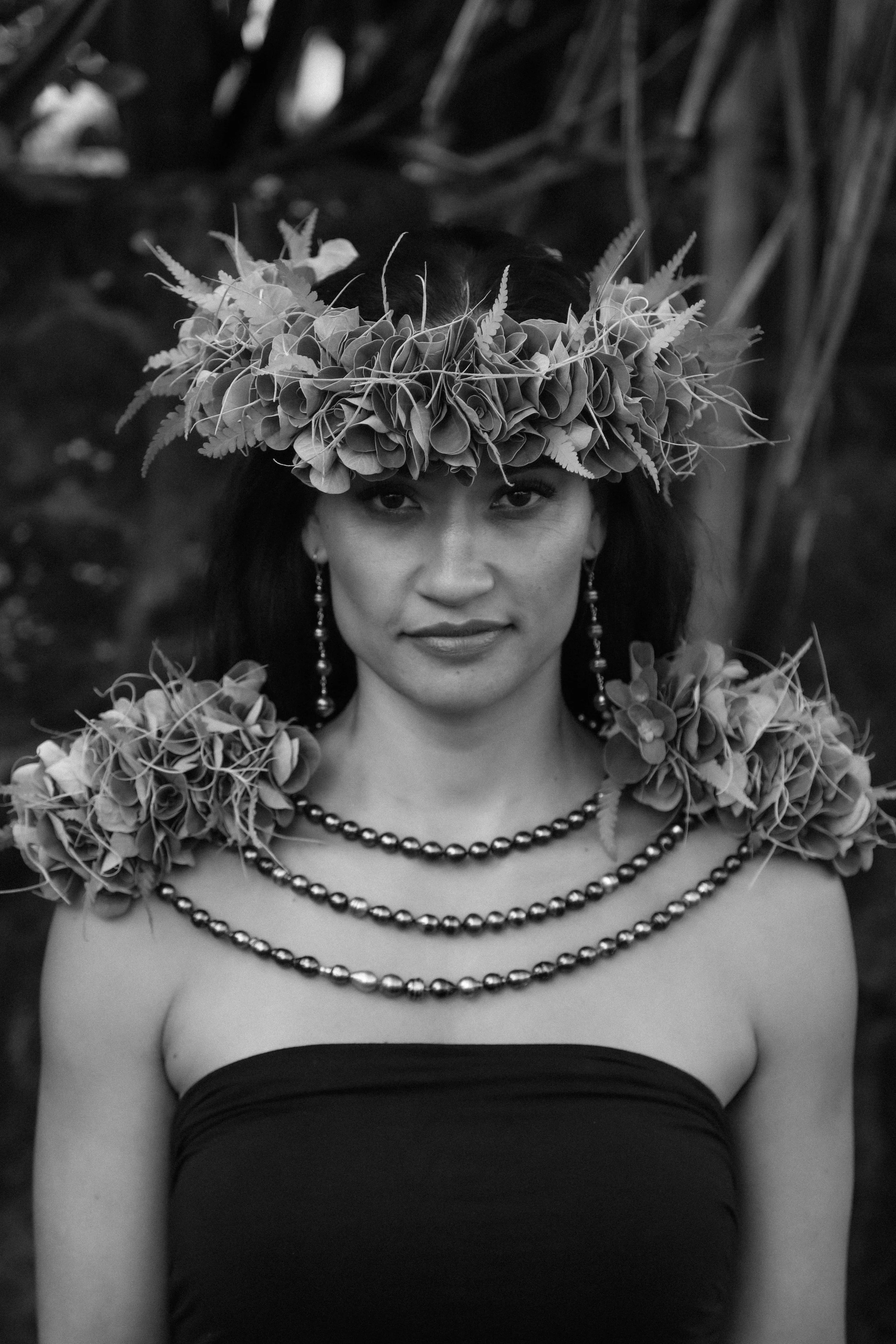 A hawaiian woman wearing a ti leaf haku, tahitian pearl earrings, and necklaces, looking directly at the camera in a black and white photo.