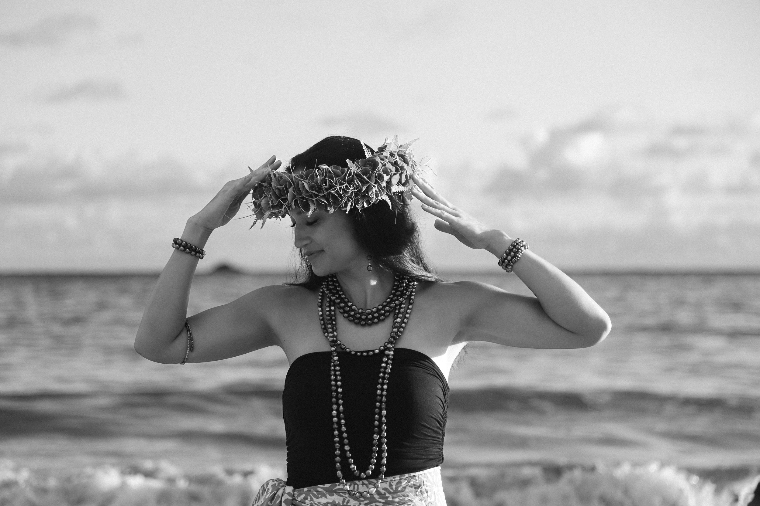 A black and white photograph of a hawaiian woman wearing a ti leaf haku and tahitian pearl necklaces stands on a beach, adjusting her haku lei while looking down with a gentle smile, dressed in a strapless top, with the ocean in the background.