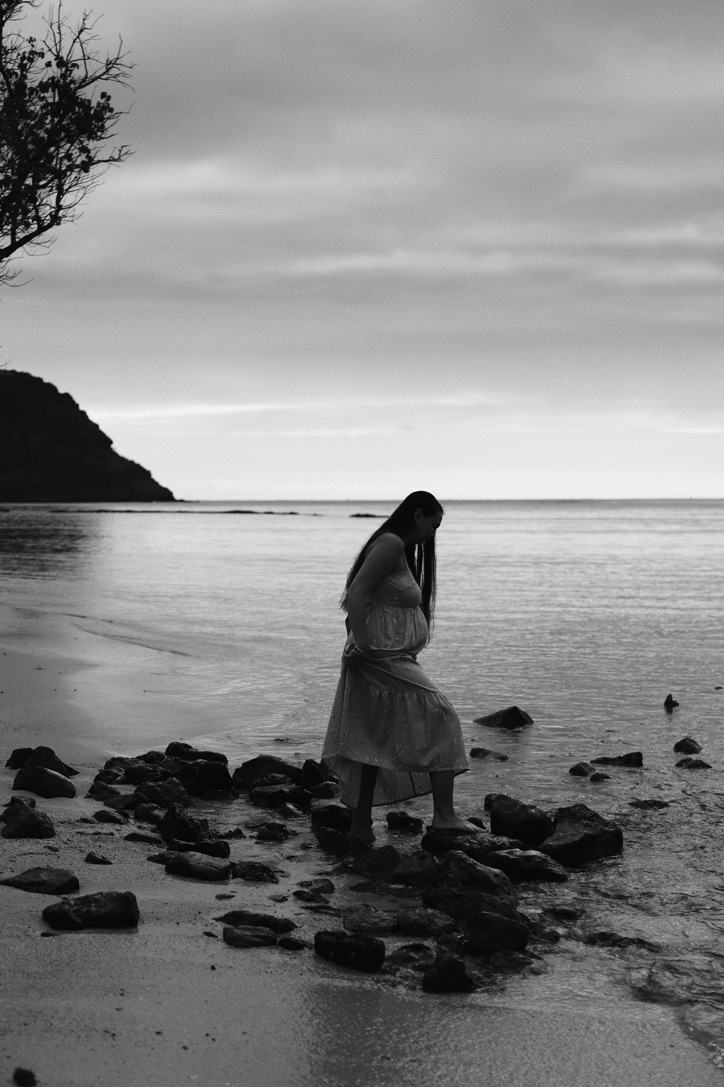 A woman in a long dress stands barefoot on rocks at the edge of a calm beach, looking down at the ground, with a distant view of a small hill or mountain.