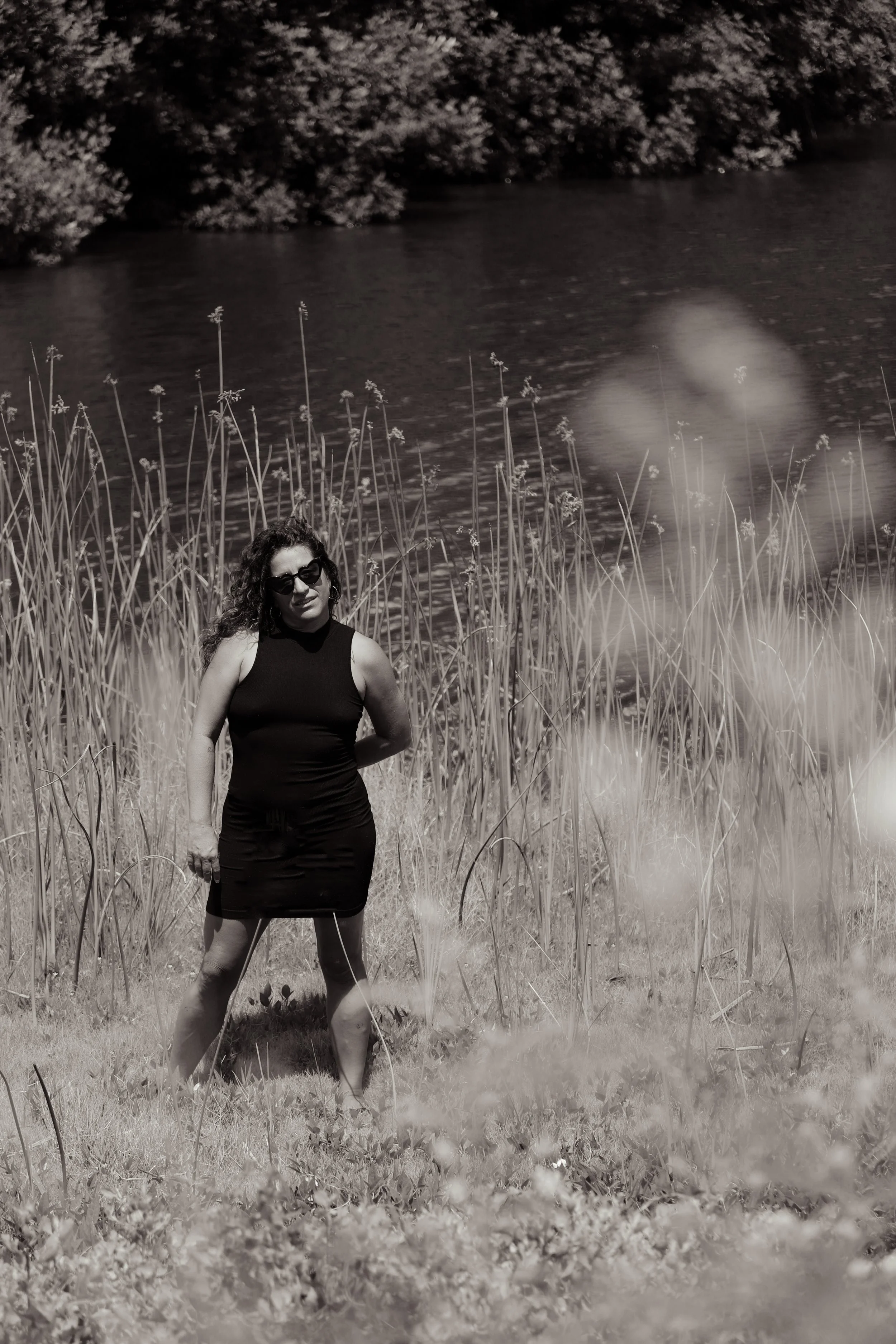 A woman in a black sleeveless dress and sunglasses standing outdoors near a body of water, with tall grass and plants around her, in black and white.