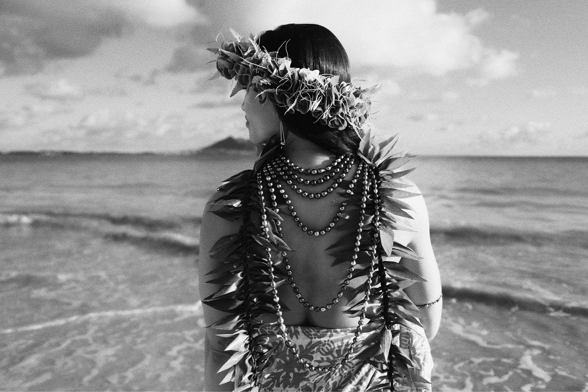 A black and white photo of a woman wearing a ti leaf haku lei, multiple tahitian pearl necklaces, and a ti leaf lei stands on a beach facing the ocean, with a cloudy sky in the background.
