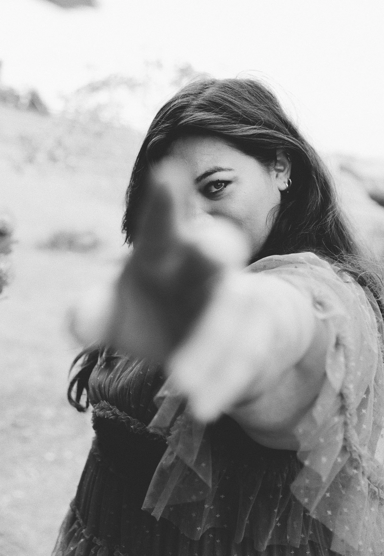 Black and white photo of a woman with long hair pointing towards the camera, with her face partially obscured by her hand, in an outdoor setting.