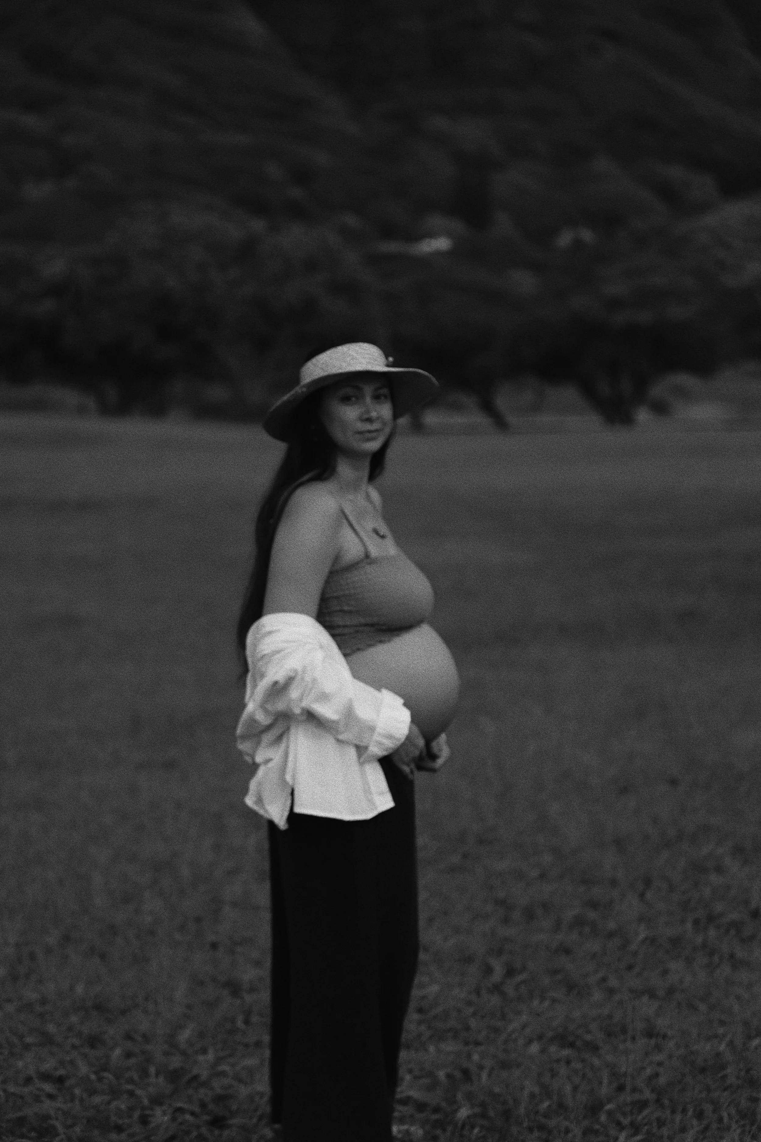 A pregnant woman standing outdoors in a field, wearing a hat and a sleeveless top, with a jacket partially draped over her arms, looking at the camera.