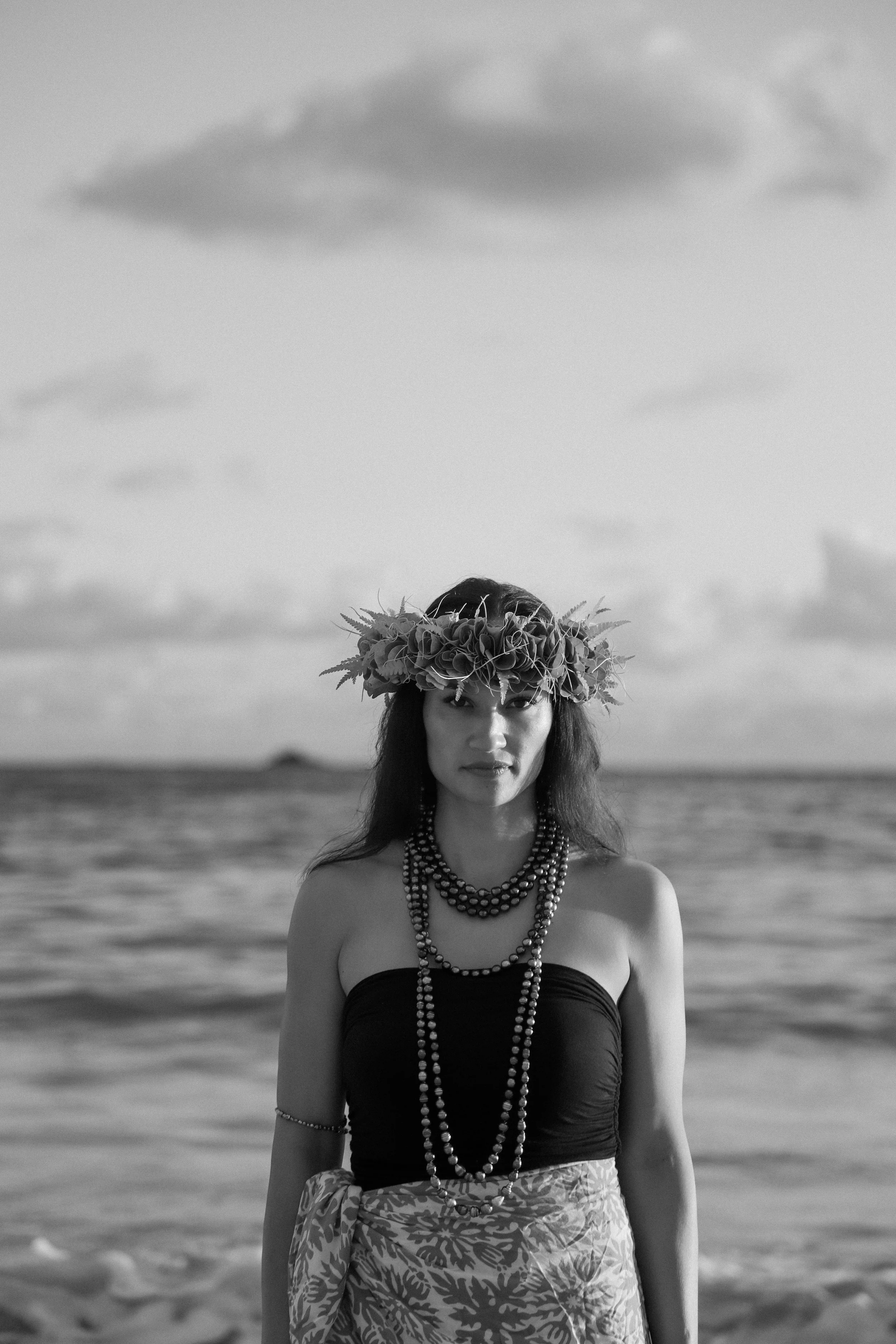 Black and white photo of a woman at the beach wearing a ti leaf haku lei, multiple tahitian pearl necklaces, and a strapless top.