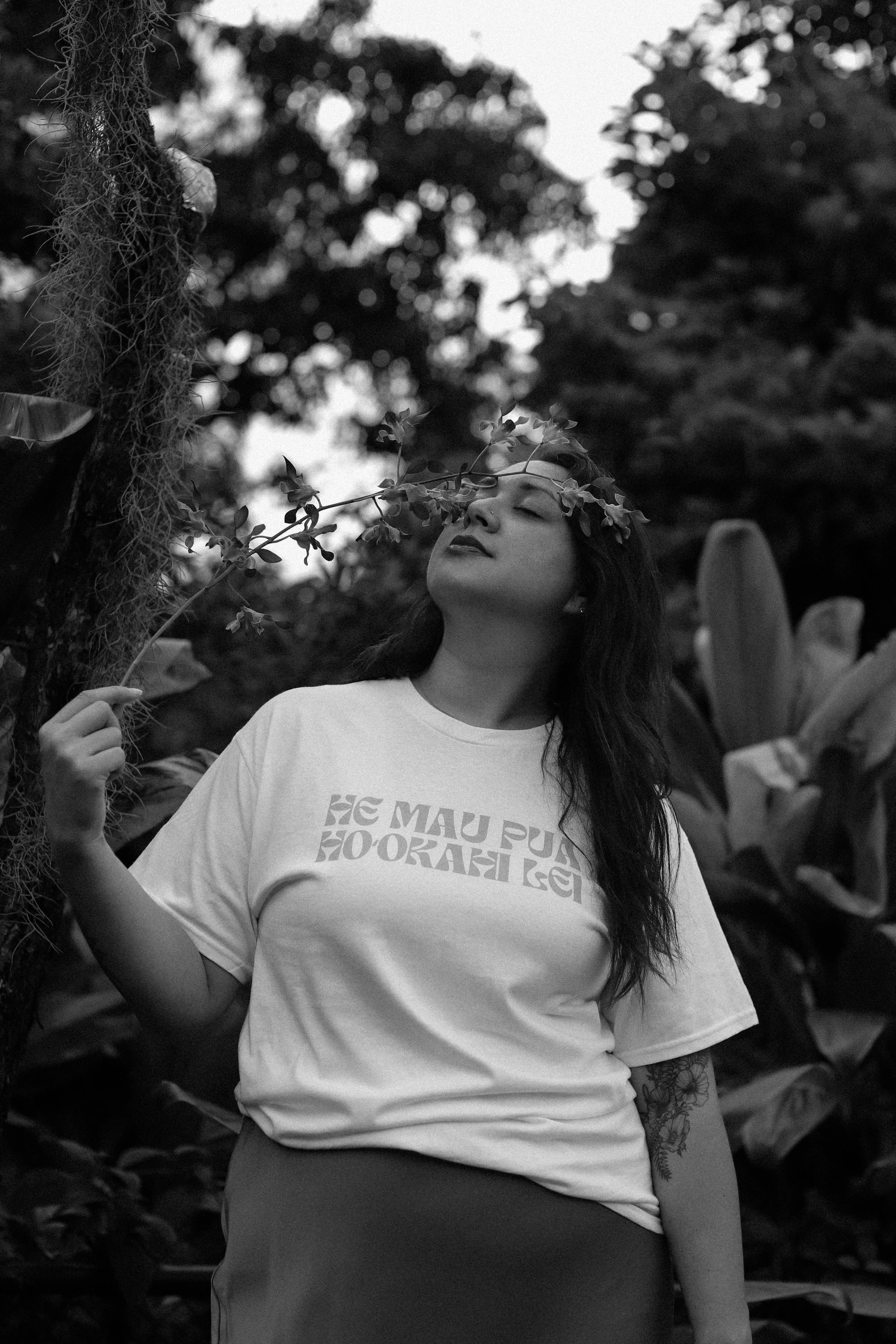 A woman with long dark hair and a floral crown on her head, standing outdoors among plants and trees, holding a branch with leaves close to her face, her eyes closed, in black and white.