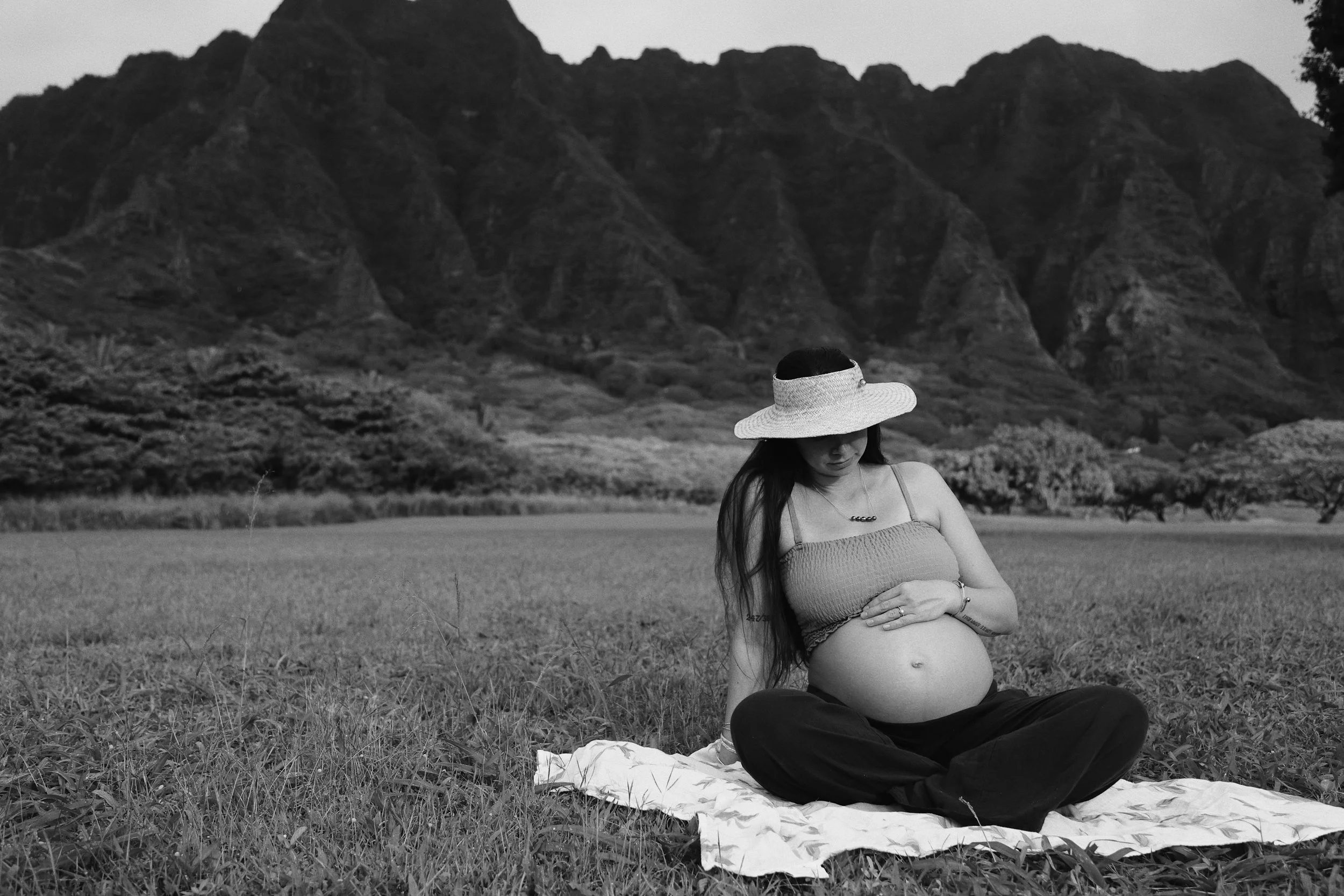 A pregnant woman sitting cross-legged on a blanket in a grassy field with mountains in the background, wearing a wide-brimmed hat and a sleeveless top.