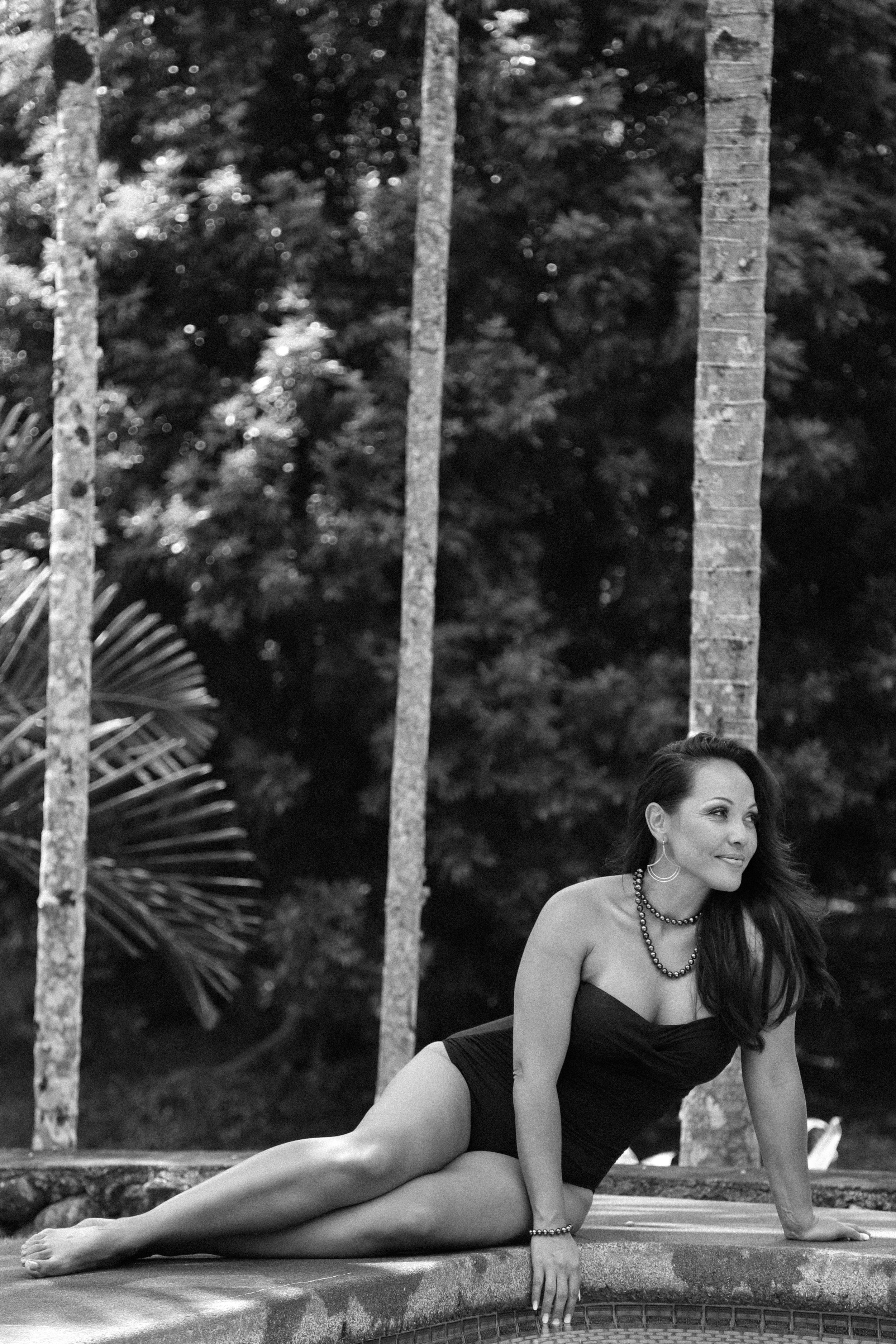 A woman in a black swimsuit relaxing on the edge of a pool, surrounded by trees and tropical plants, in black and white photo.