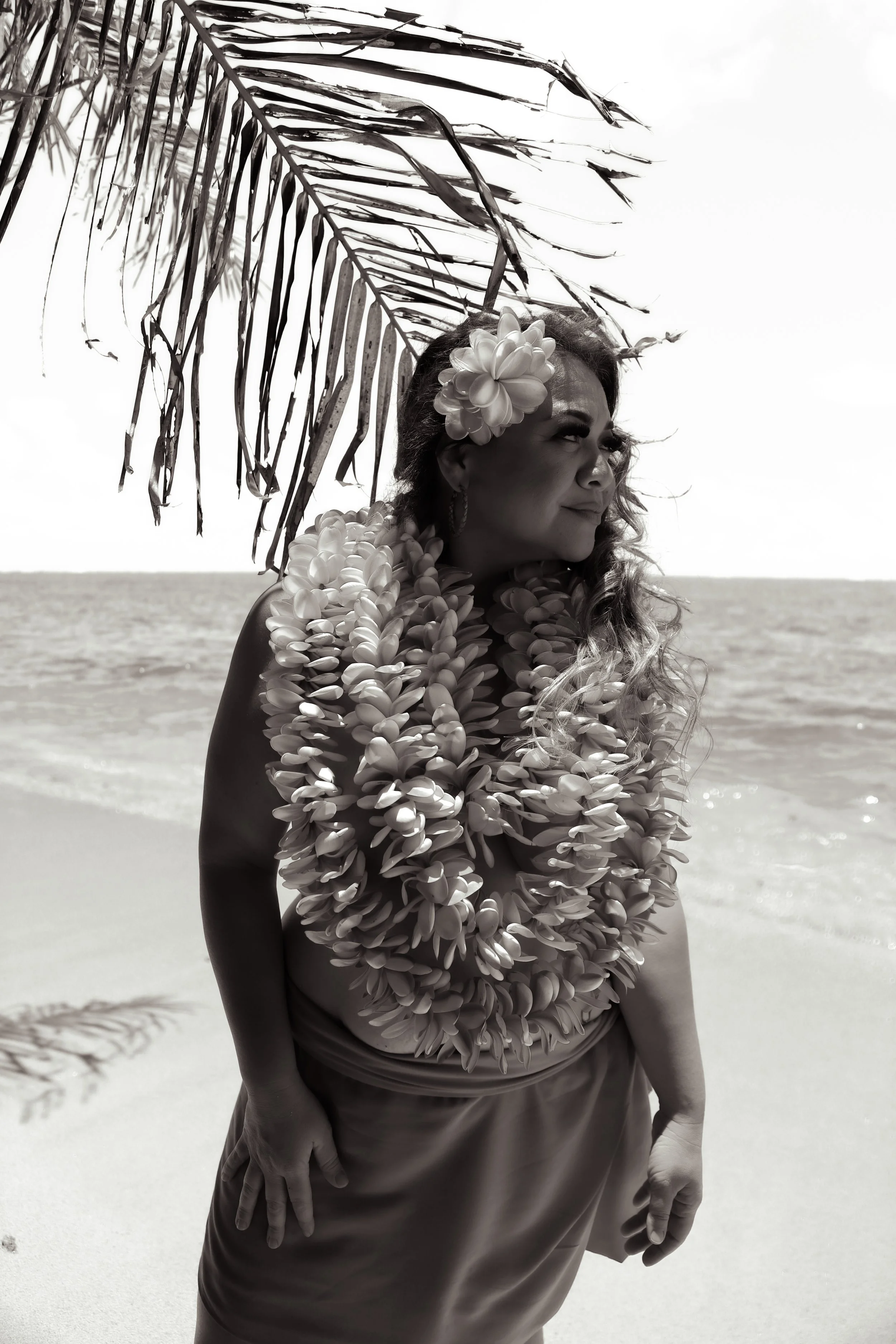 A woman standing on a beach under a palm tree, wearing a floral lei and hair accessory, looking into the distance, with the ocean in the background.