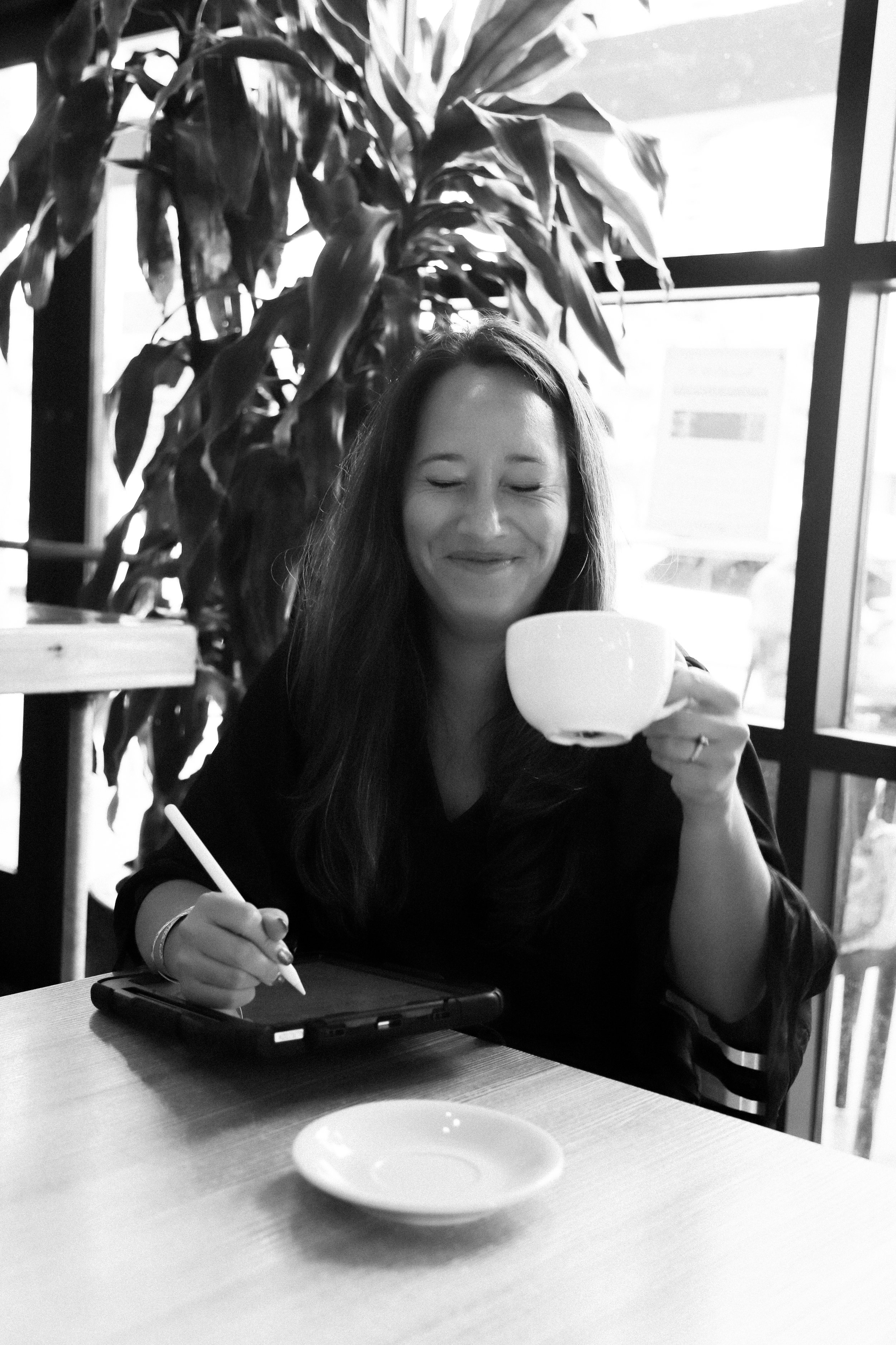 A woman with long hair smiling with her eyes closed, holding a teacup in her right hand, and writing on a tablet with her left hand, sitting at a table in a cafe with large windows and indoor plants.
