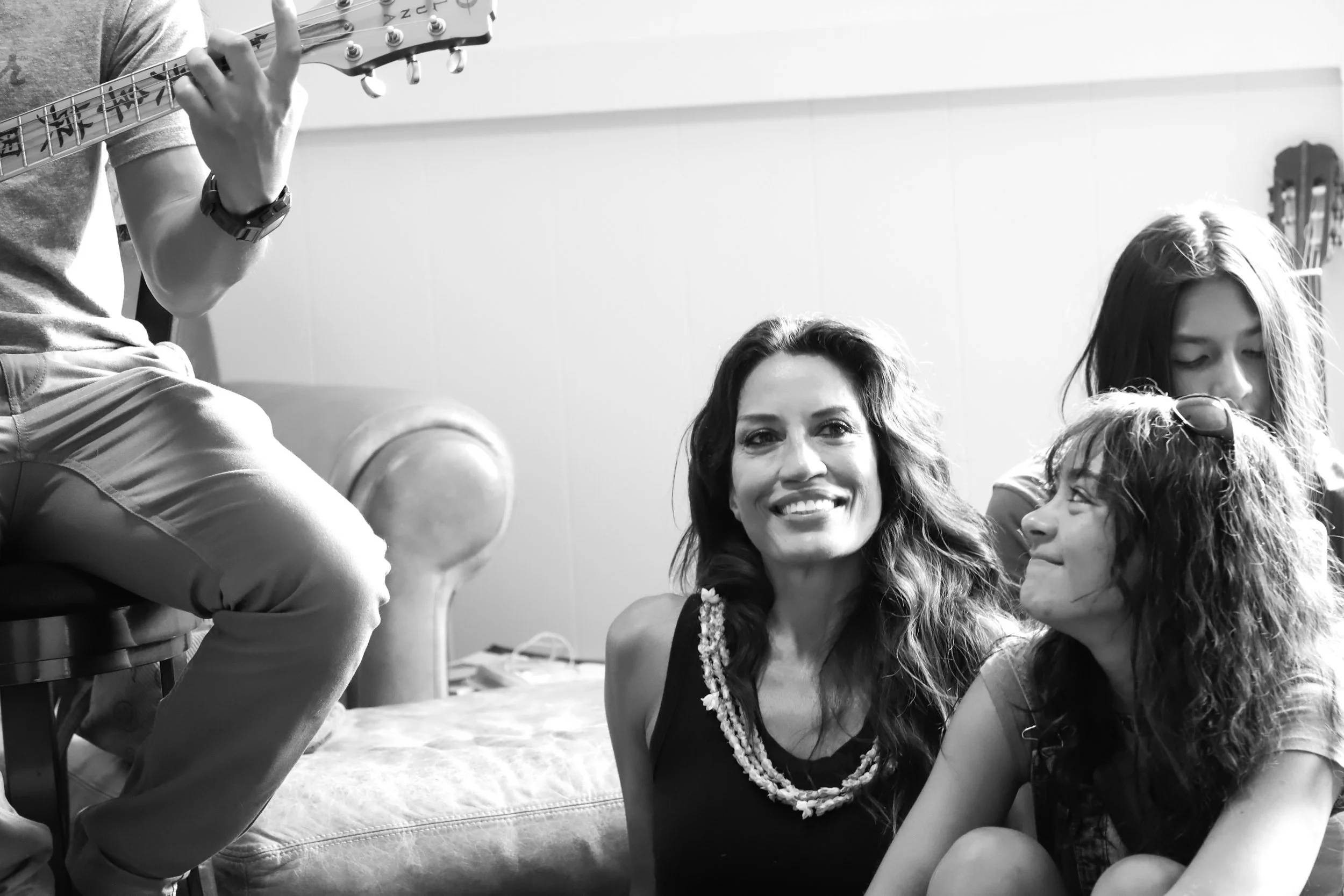 Four women sitting together, smiling and enjoying a moment indoors, with one woman playing a guitar.