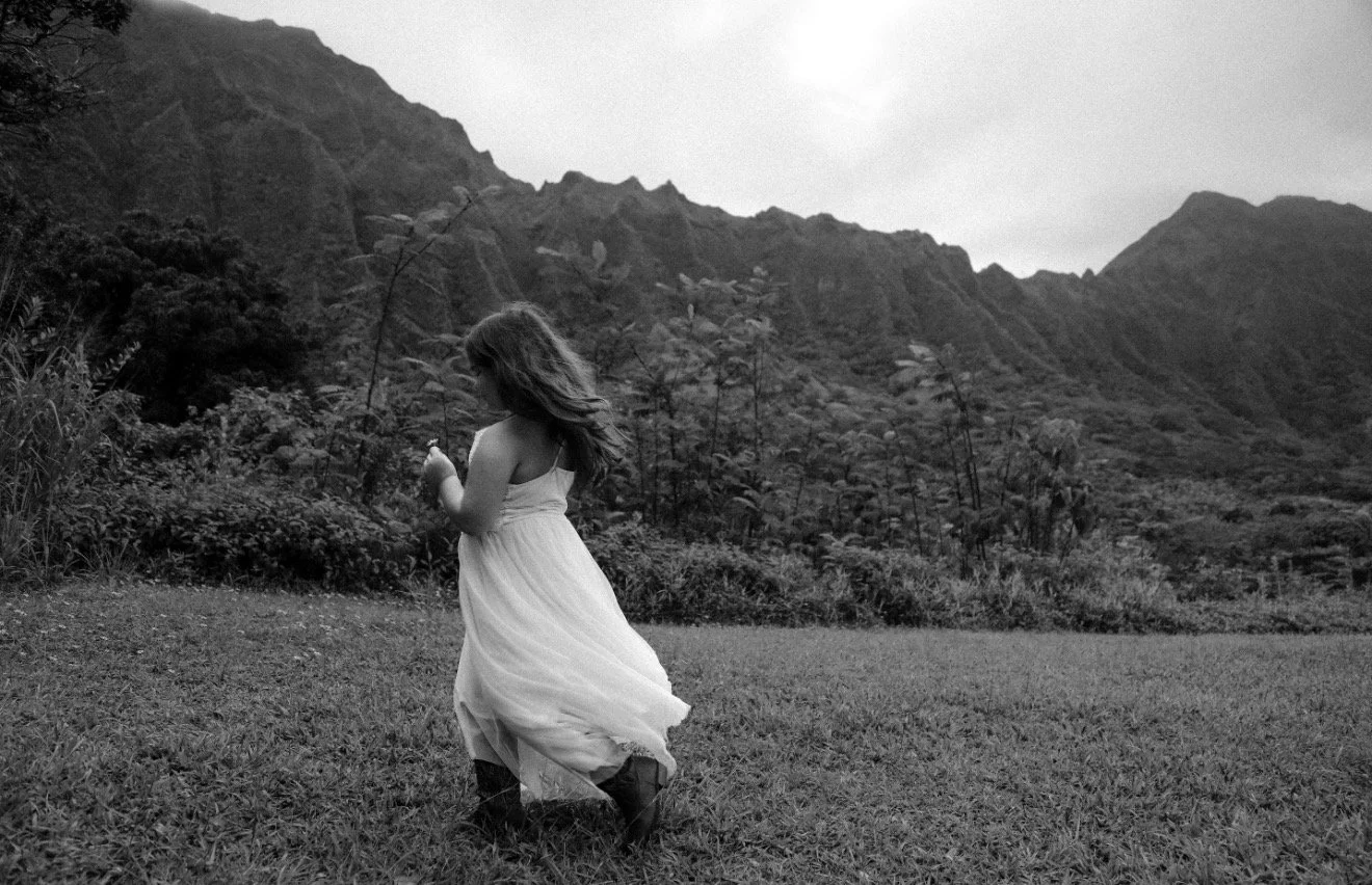 A girl in a white dress walking on grass with mountains in the background, black and white photo.
