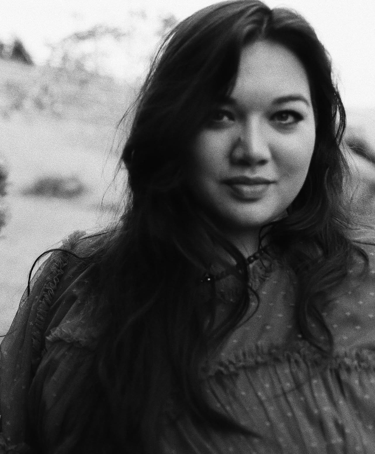 A black and white portrait of a woman with long dark hair, smiling softly, outdoors with a blurred natural landscape in the background.