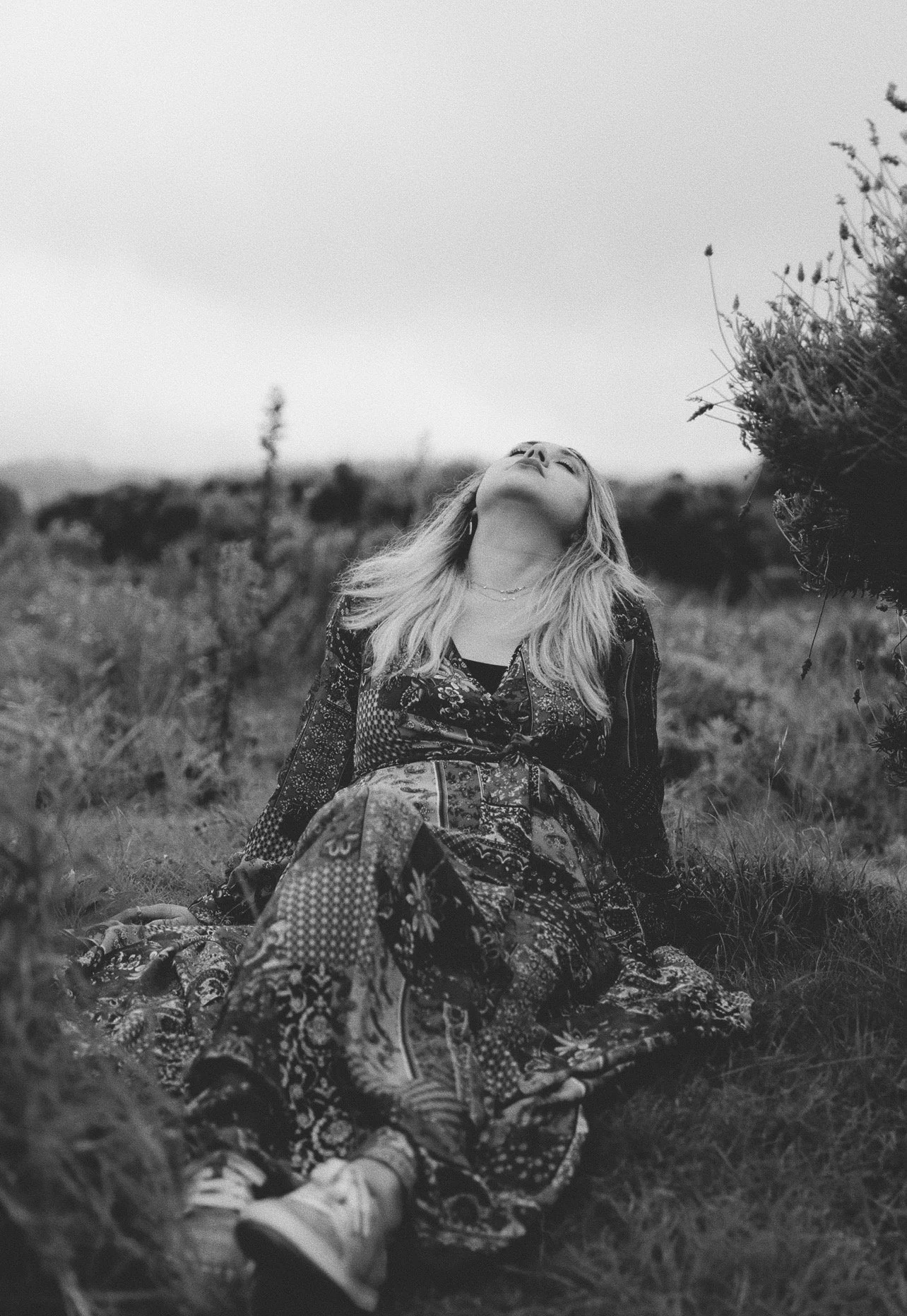 A woman with long blonde hair sitting on grass in a field, looking up with her eyes closed, captured in black-and-white.