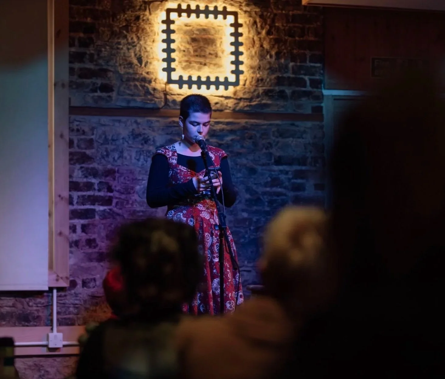 A young woman with short hair performing on stage, standing in front of a microphone, reading from a phone or paper. She is wearing a patterned dress and is illuminated by stage lighting. The background features a brick wall and a decorative light fixture.