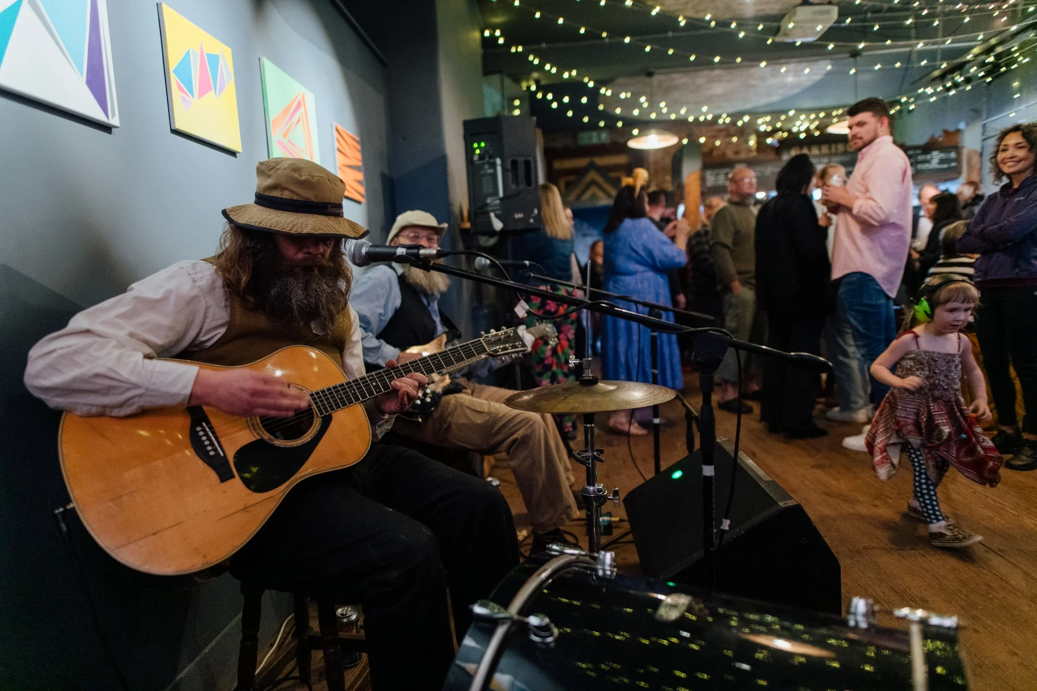 Musicians performing live in a lively venue with colorful abstract art on the wall and string lights overhead. A man with a beard and hat is playing an acoustic guitar, and another musician with glasses and a hat is playing drums. The audience, inclu