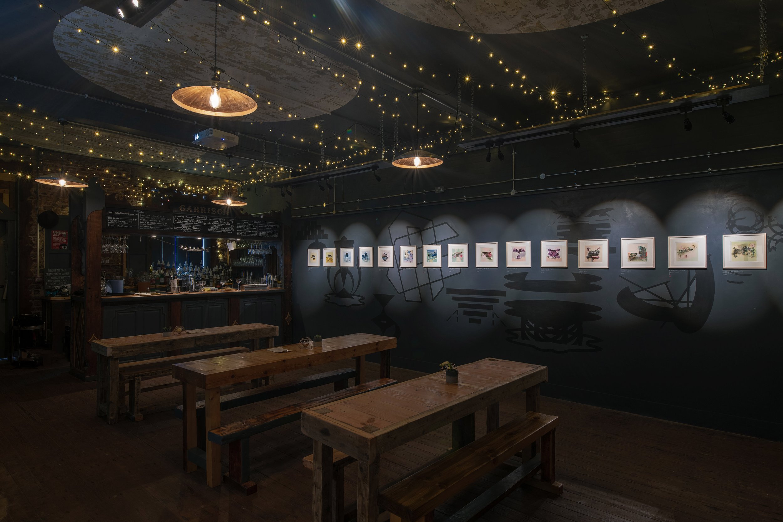 Interior of a dimly lit café or bar with wooden tables and benches, string lights on the ceiling, framed artwork on the dark wall, and a small bar area with glasses and bottles.