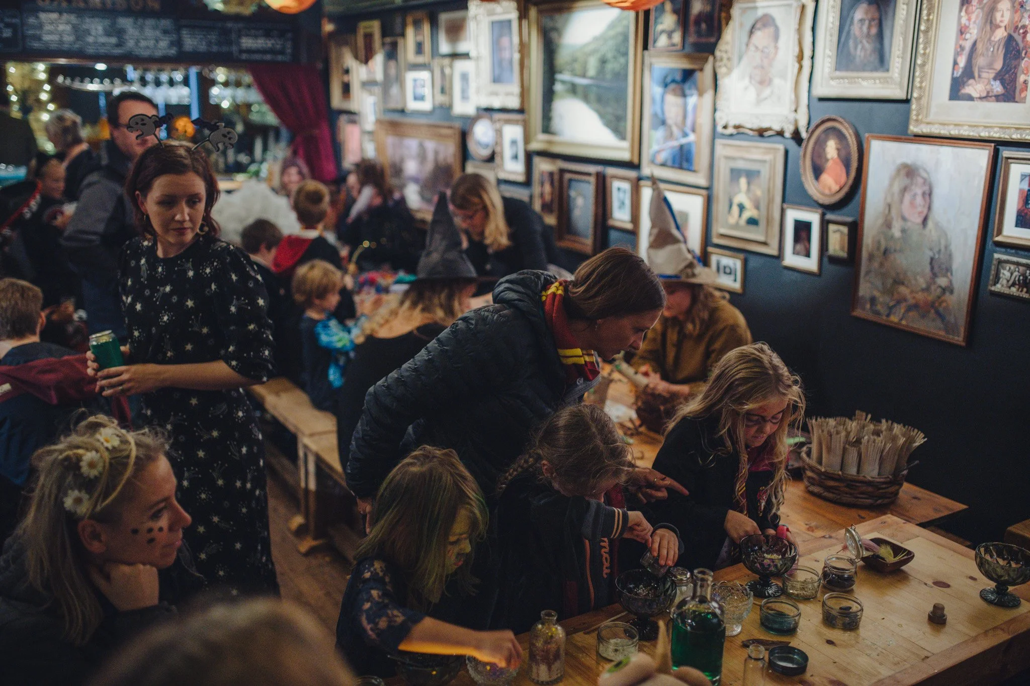 People gathered at a craft or art event in a gallery, with walls decorated with many framed paintings and photographs, children and adults participating and observing.