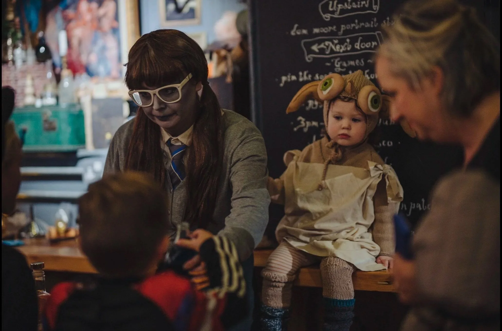 Kids and adults at a restaurant, one girl dressed in a spooky costume with face paint and glasses, and a small child in a costume with a hat resembling a rabbit or other animal, sitting at a table.