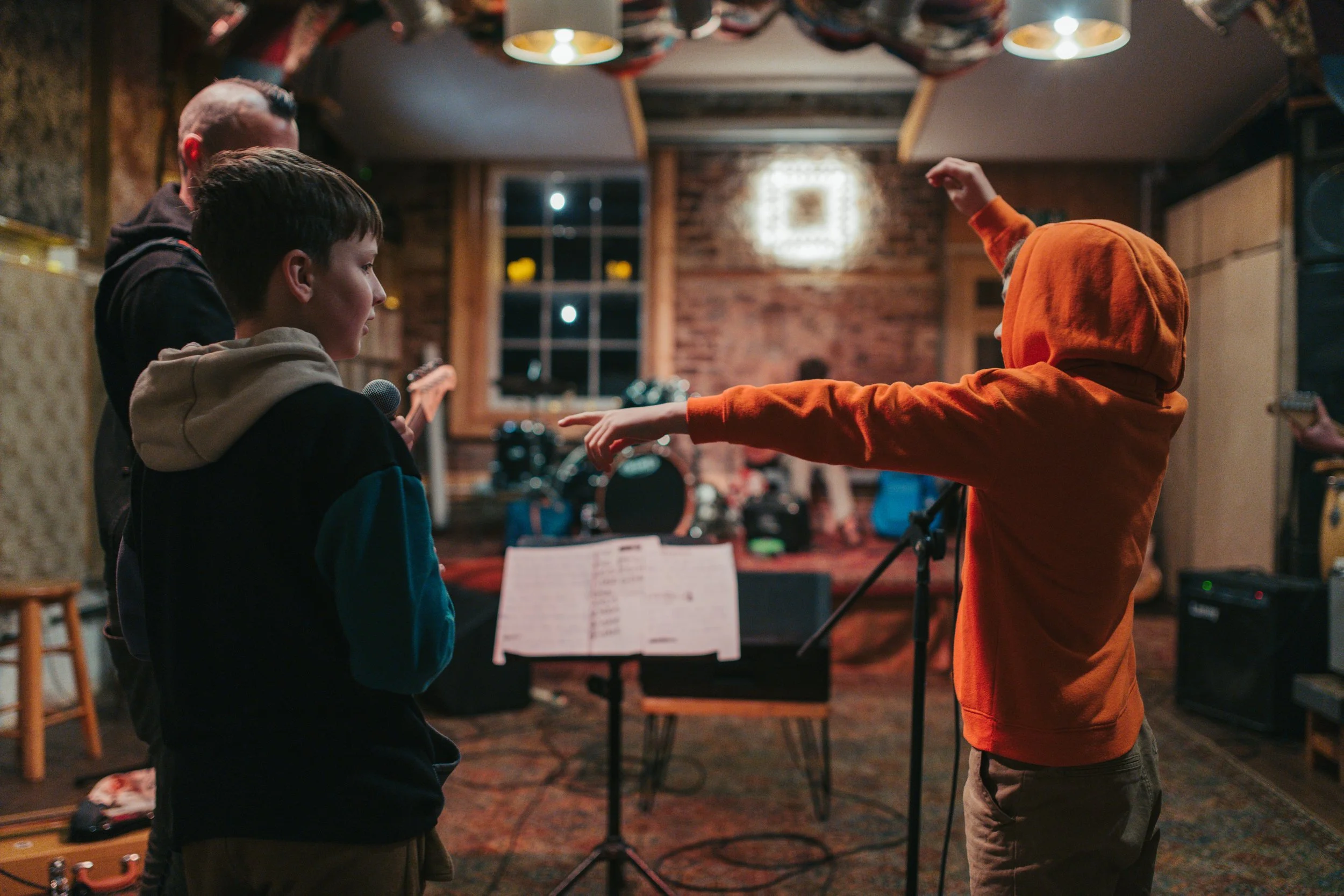 Three children in a music rehearsal room, one in an orange hoodie with hood up gesturing while another holds a microphone and a third looks on.