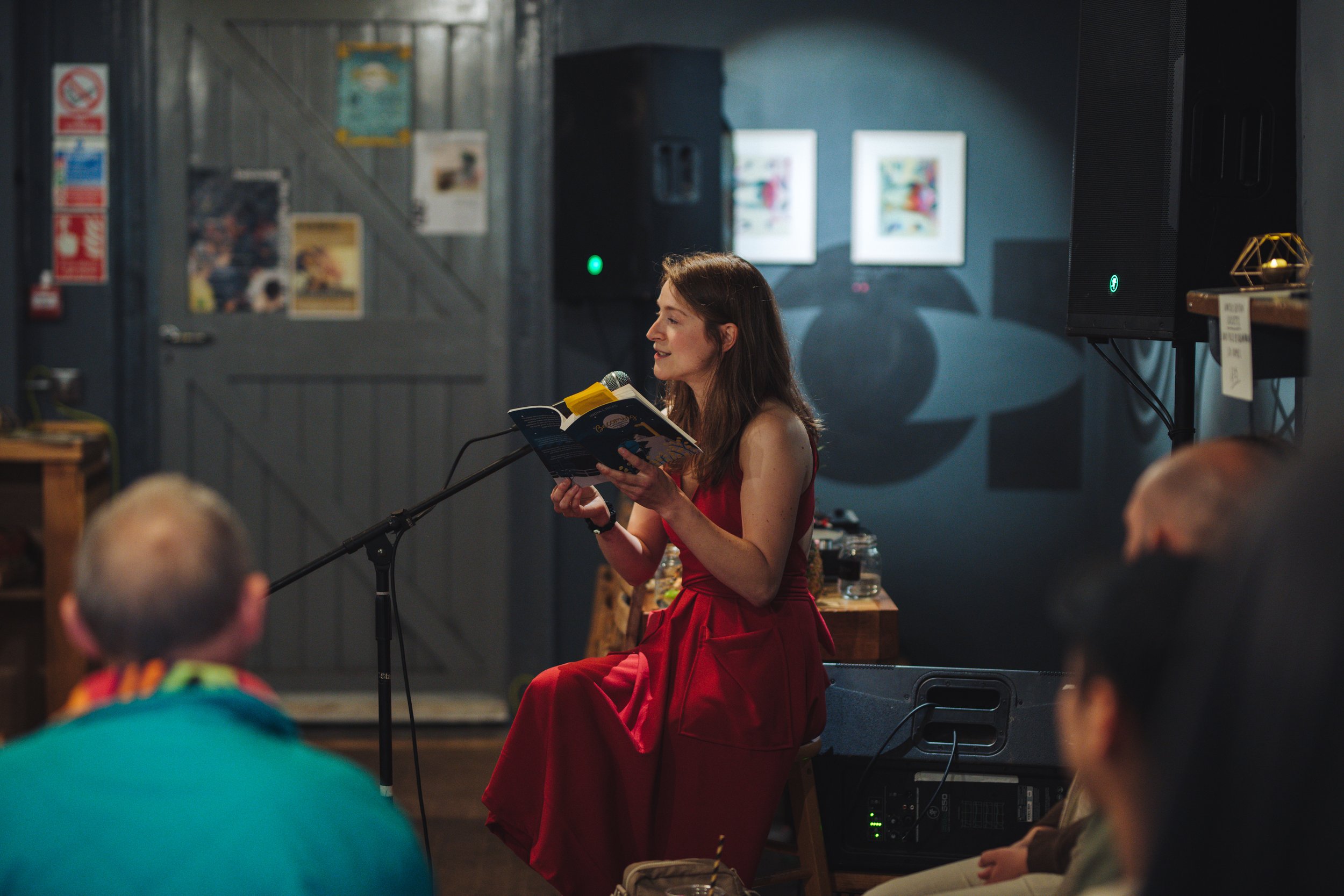 A woman in a red dress reading from a book into a microphone while sitting on a stool at a small event, with an audience in front of her, in an indoor setting with artwork on the wall behind her.