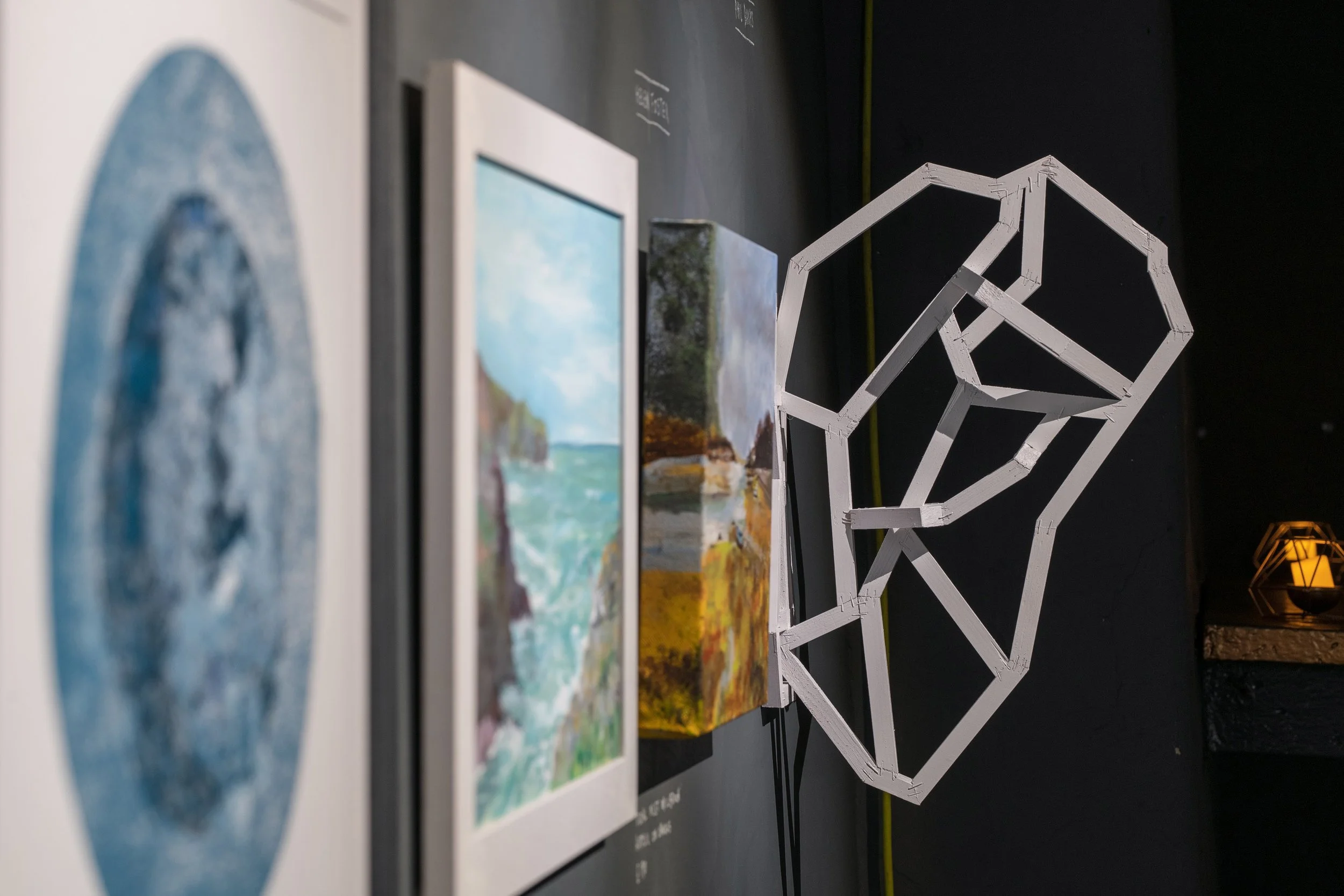 Gallery display with framed artwork and a geometric white sculpture against a black wall, illuminated by warm lighting.