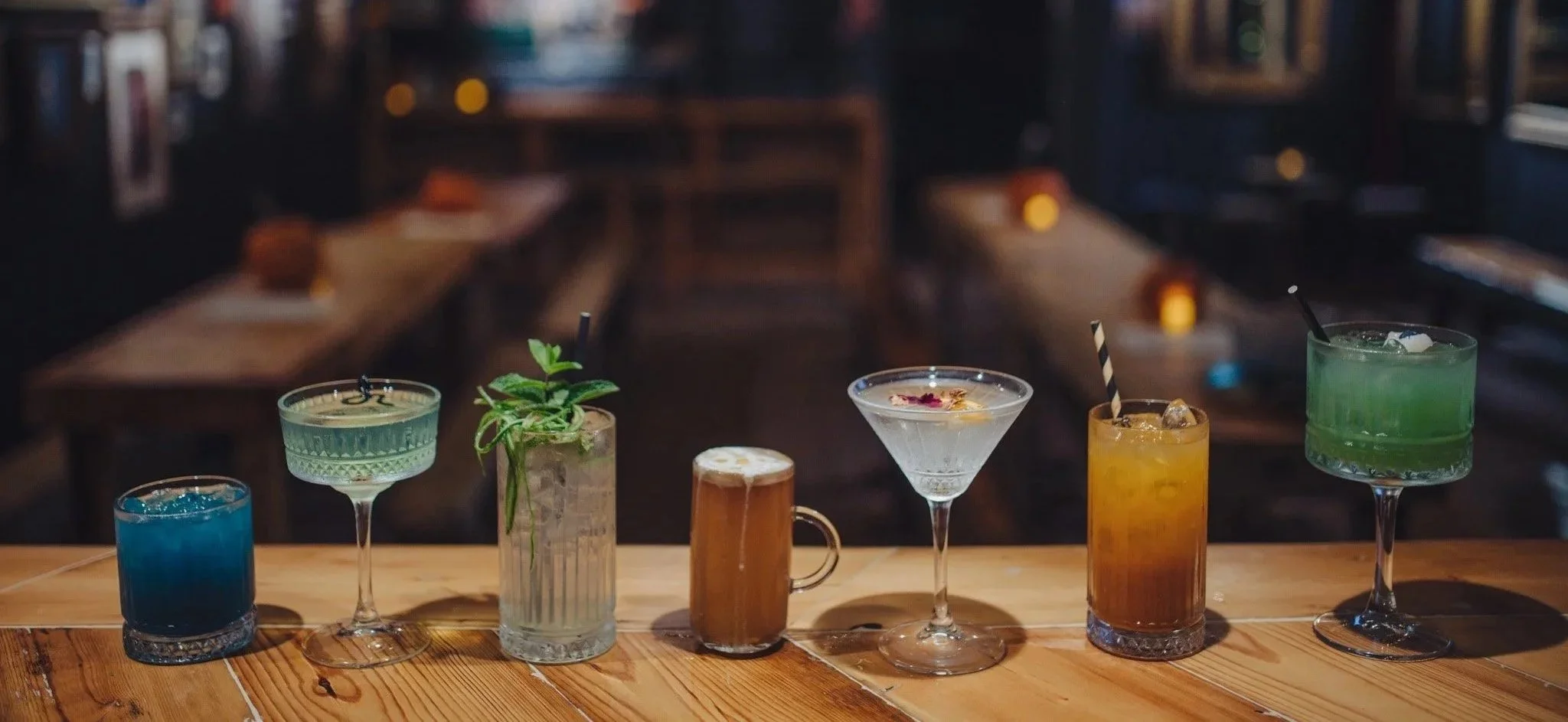 A row of seven colorful cocktails on a wooden bar counter, with a blurred bar or restaurant interior in the background.