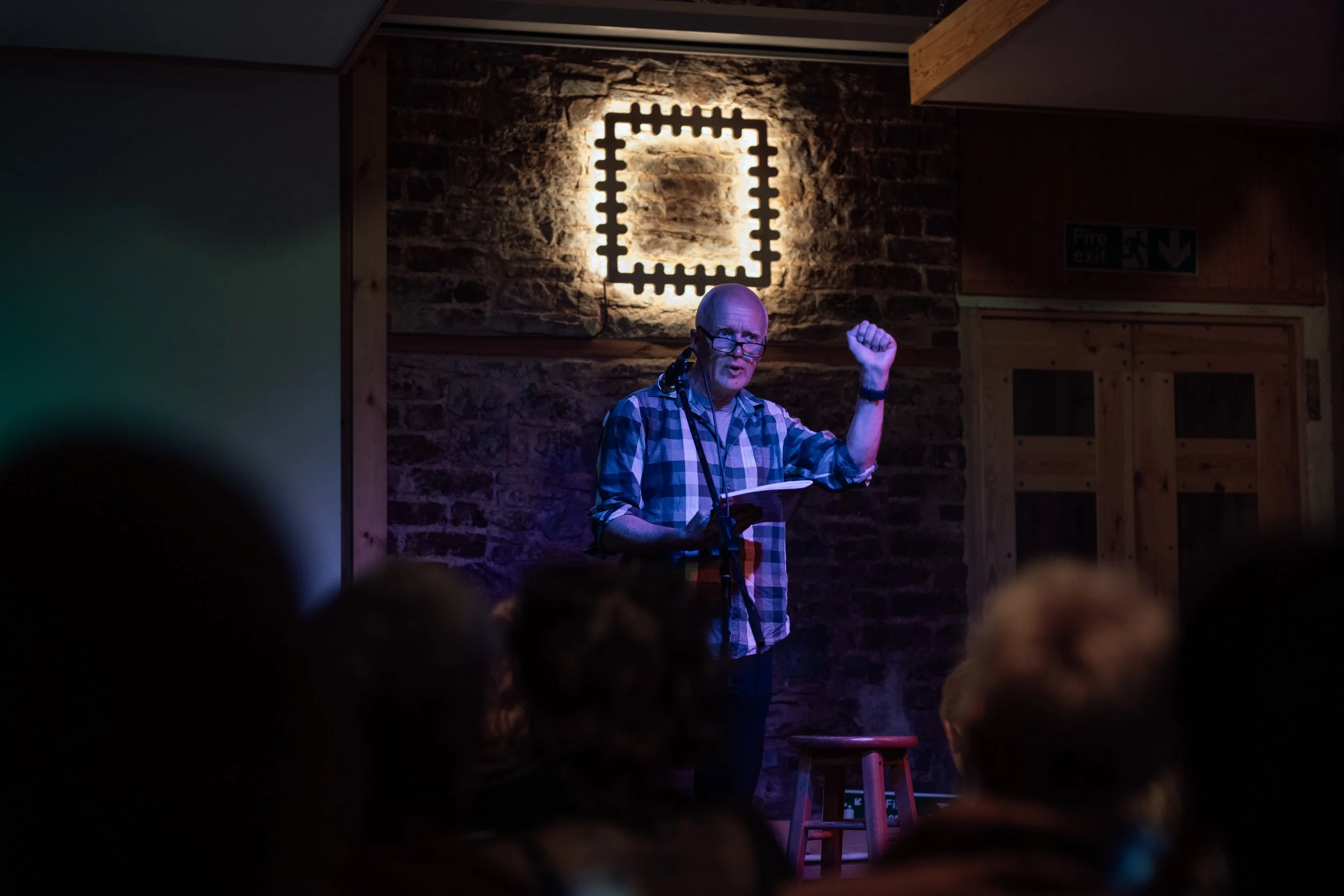 A man with glasses and a plaid shirt giving a presentation on stage, with a brick wall and illuminated artwork behind him, in a dimly lit venue.