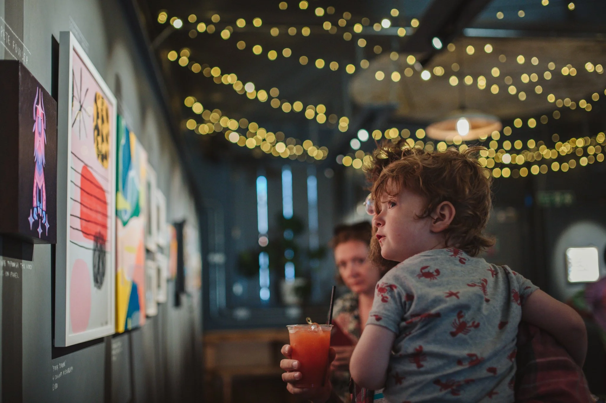 Young child with curly hair wearing a lobster-patterned shirt sitting at a table with a woman, holding a red drink, at an art gallery or cafe decorated with string lights.