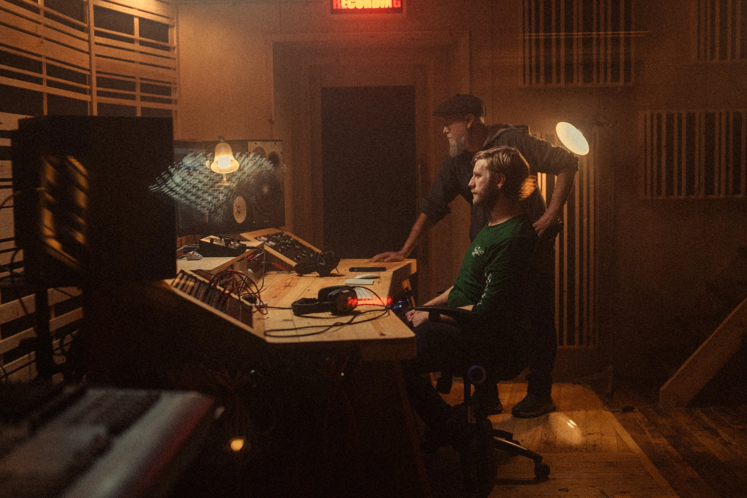 Two men in a recording studio. One is sitting at a desk with audio equipment, wearing a green shirt. The other man is standing, leaning on the desk, wearing a dark jacket and cap. The studio has wooden walls and a large speaker in the foreground.