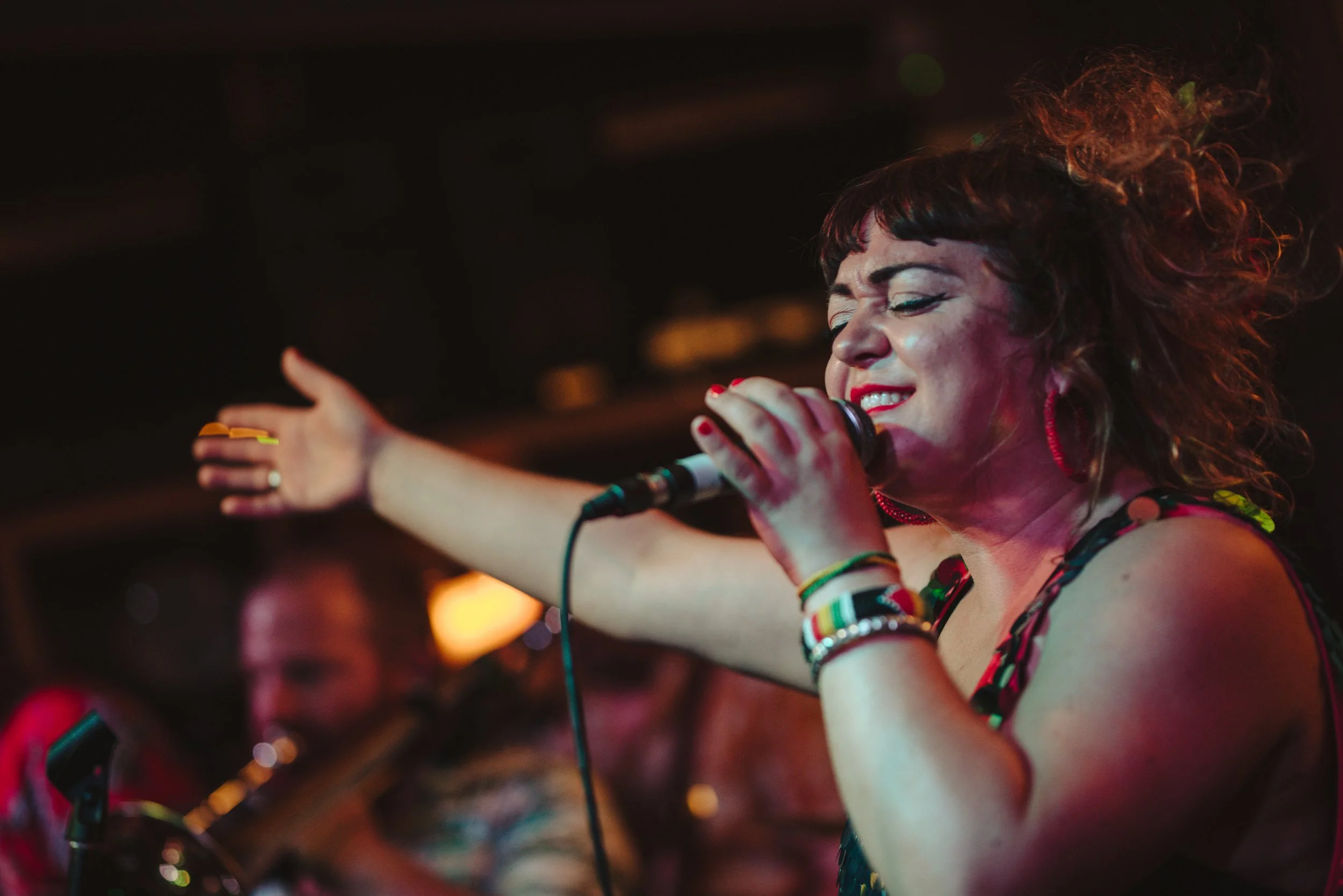 A woman singing into a microphone with her eyes closed, wearing colorful bracelets and earrings, with her arm raised in a nightclub or concert setting.