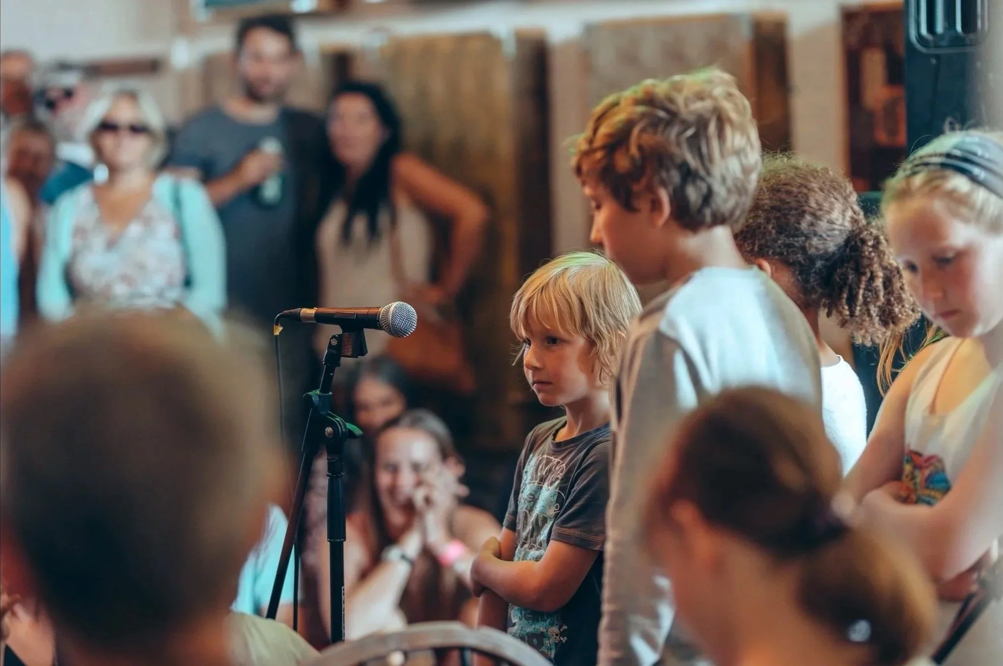 Children standing in front of microphone at a gathering or event, with audience and adults in the background.