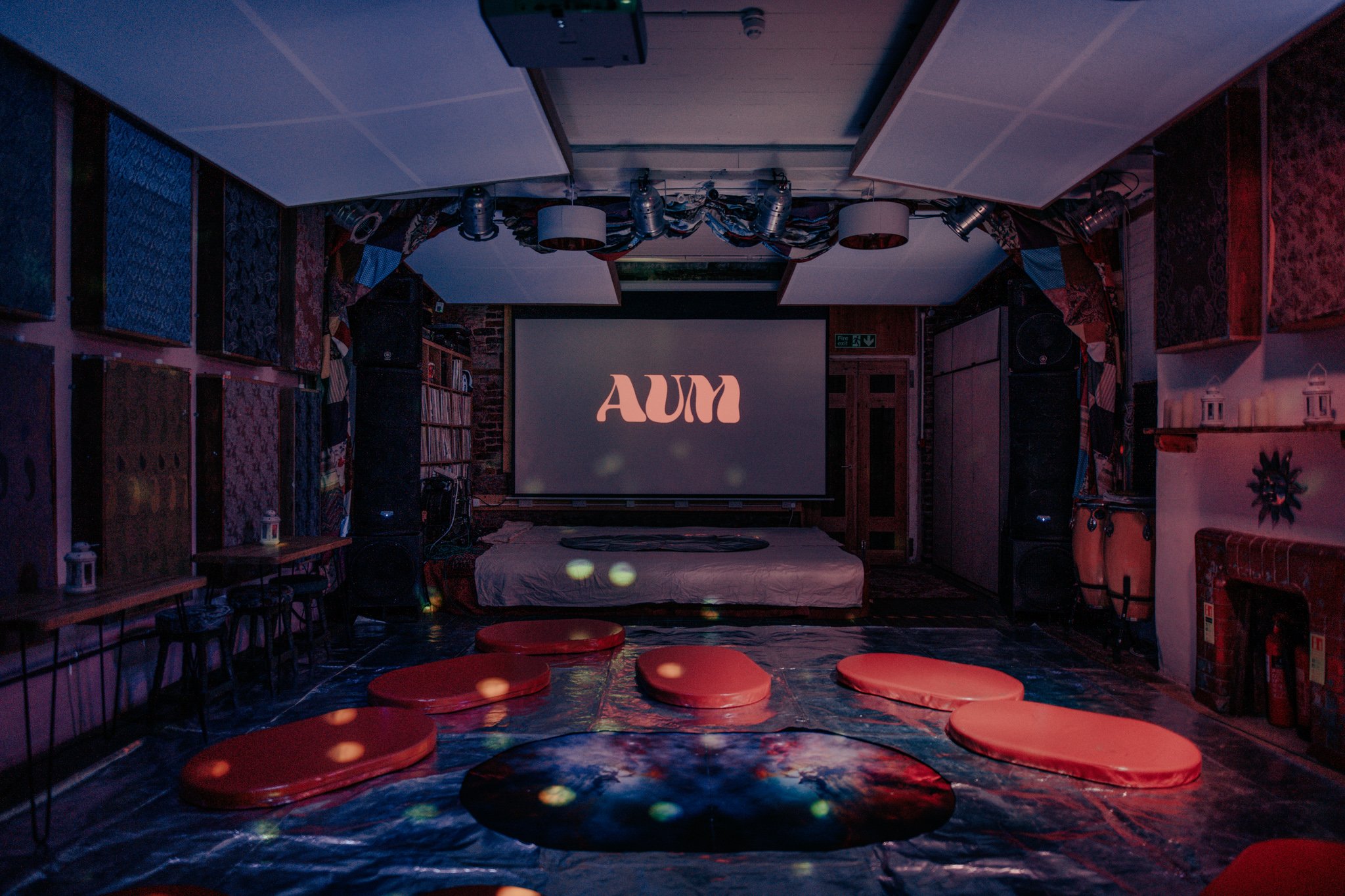 A dimly lit room with a large screen displaying the word 'AUM' in stylized font, surrounded by red cushions on the floor, and decorated with patterned fabrics, lanterns, and a fireplace.
