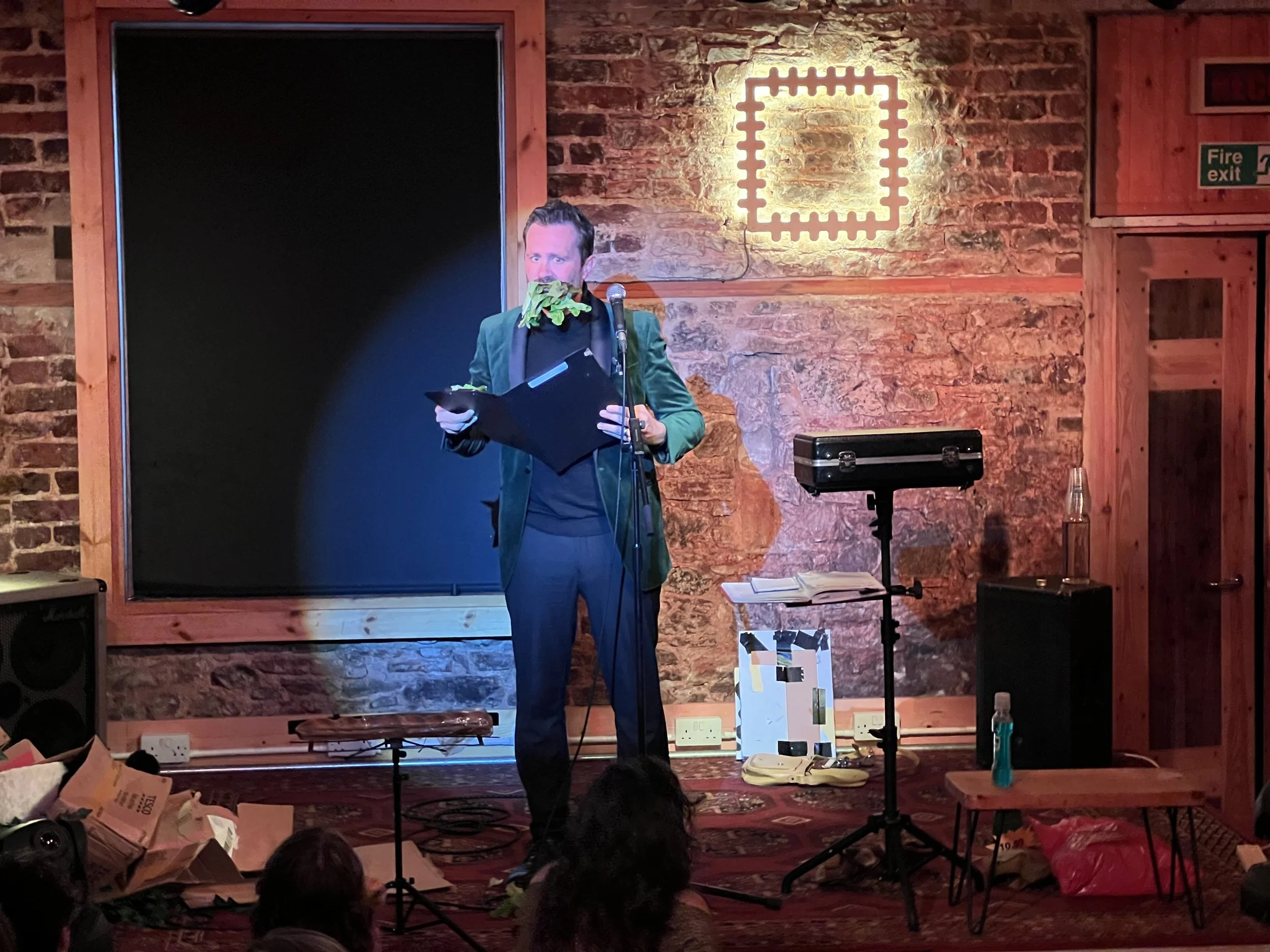 A man on stage performing with leafy props in front of his face, holding a binder and standing in front of a microphone, in a cozy venue with brick walls and soft lighting.