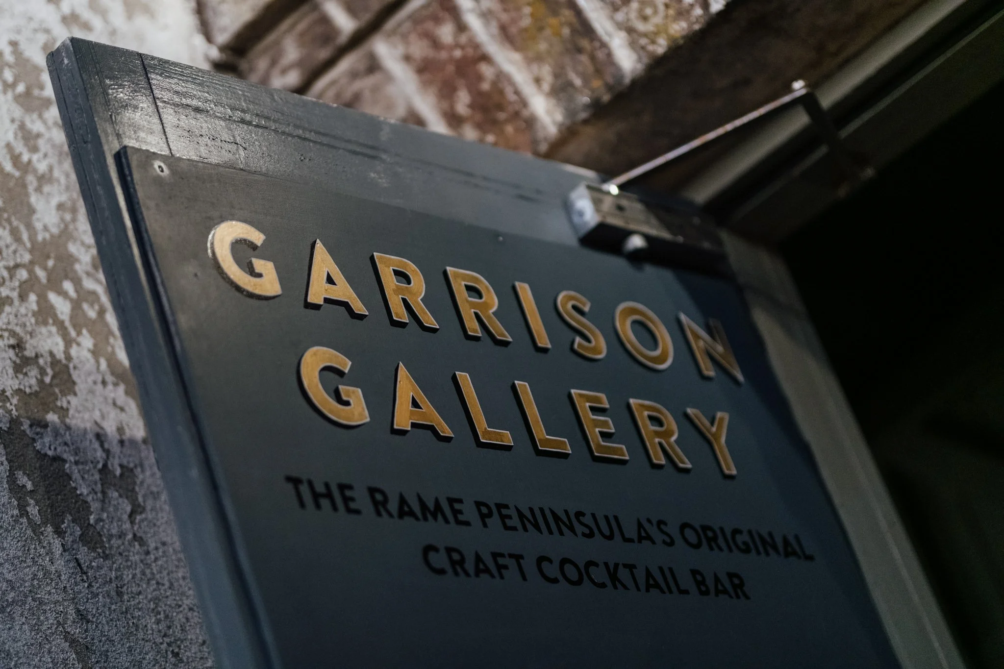 Close-up of a sign for Garrison Gallery, located at The Rame Peninsulas Original Craft Cocktail Bar, mounted on a brick wall.