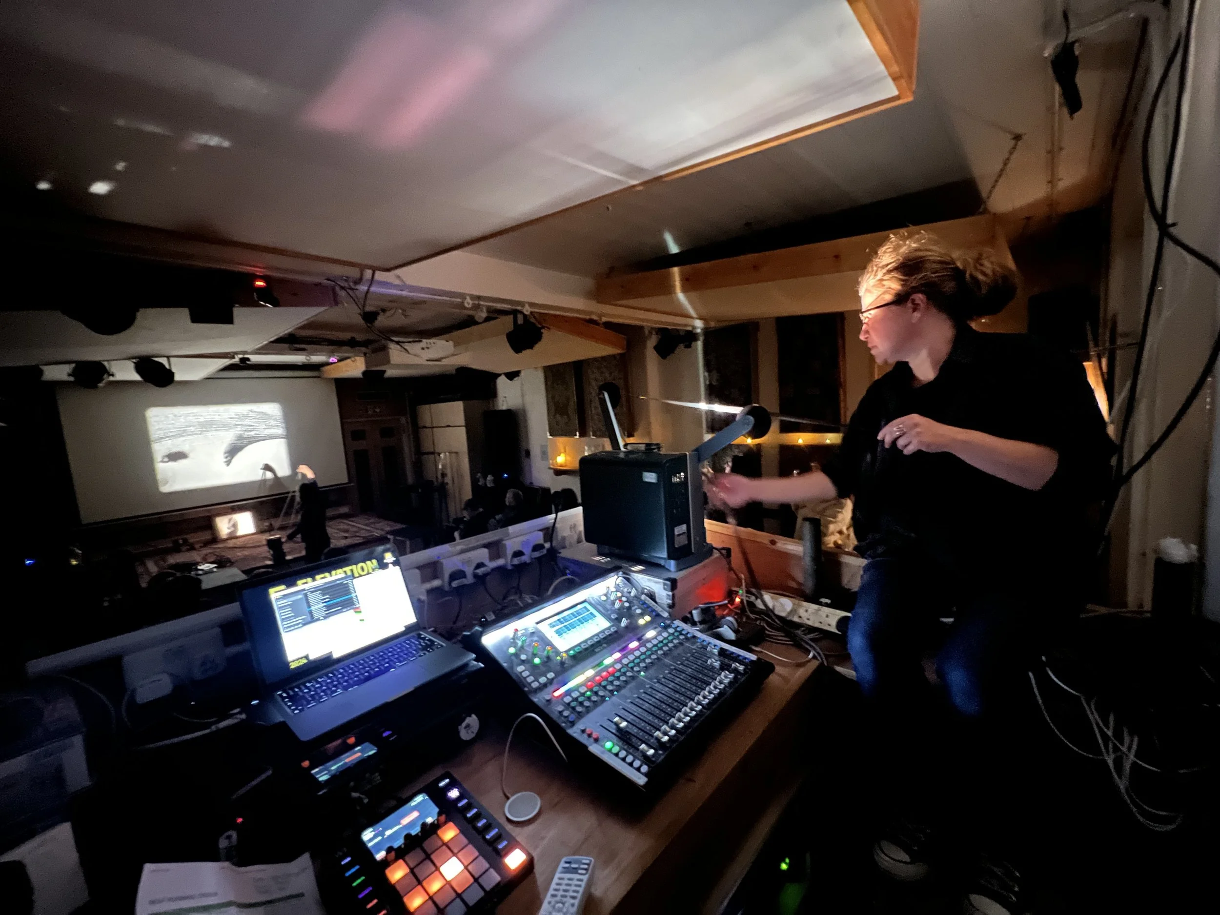 A woman with blonde hair and glasses working with DJ equipment on a platform overlooking a darkened room with a large projection screen displaying black-and-white footage.