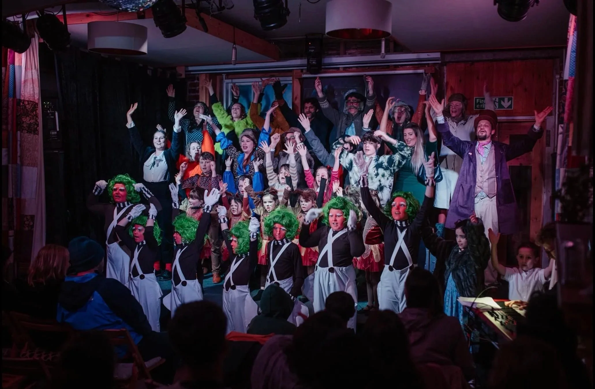 A lively stage performance with a diverse group of children and adults in colorful costumes, including some with green wigs and face paint, performing in front of an audience.