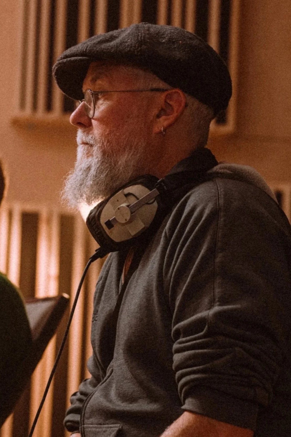 A bearded man wearing glasses, a black beret, and a dark hoodie, with headphones around his neck, in a recording studio.