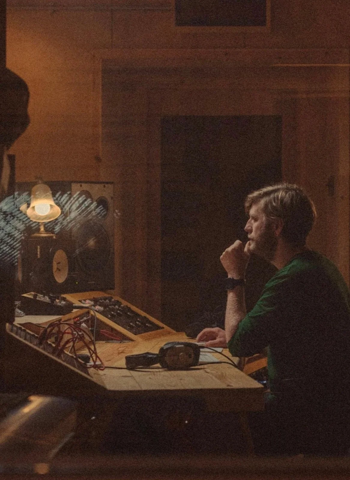 A man sitting in a recording studio with wood-paneled walls, resting his chin on his hand, looking at a mixing console, with a speaker and a small lamp in the background.