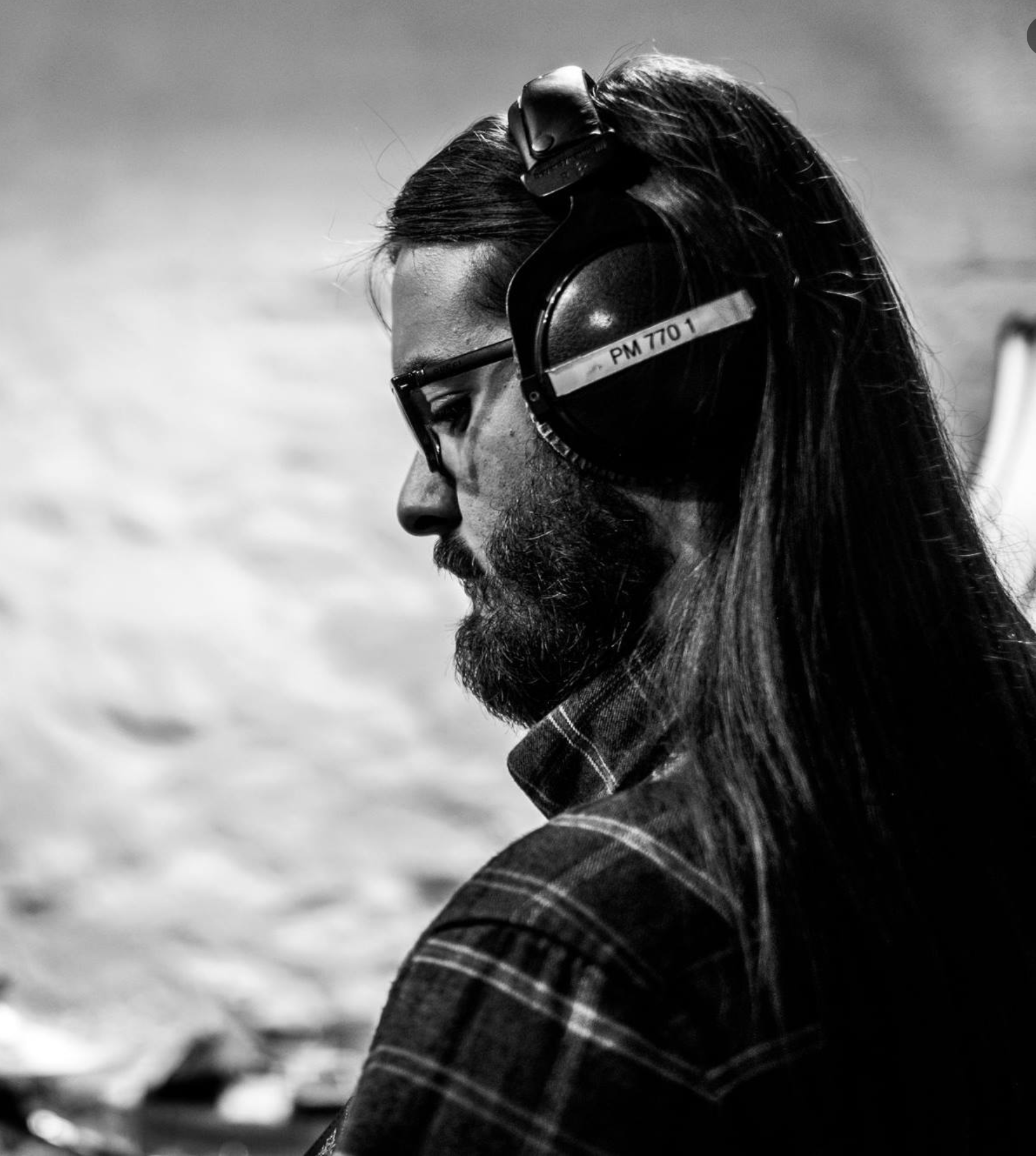 A man with long hair and a beard, wearing glasses and large headphones, is looking down, captured in profile. The background appears to be a blurry outdoor setting.