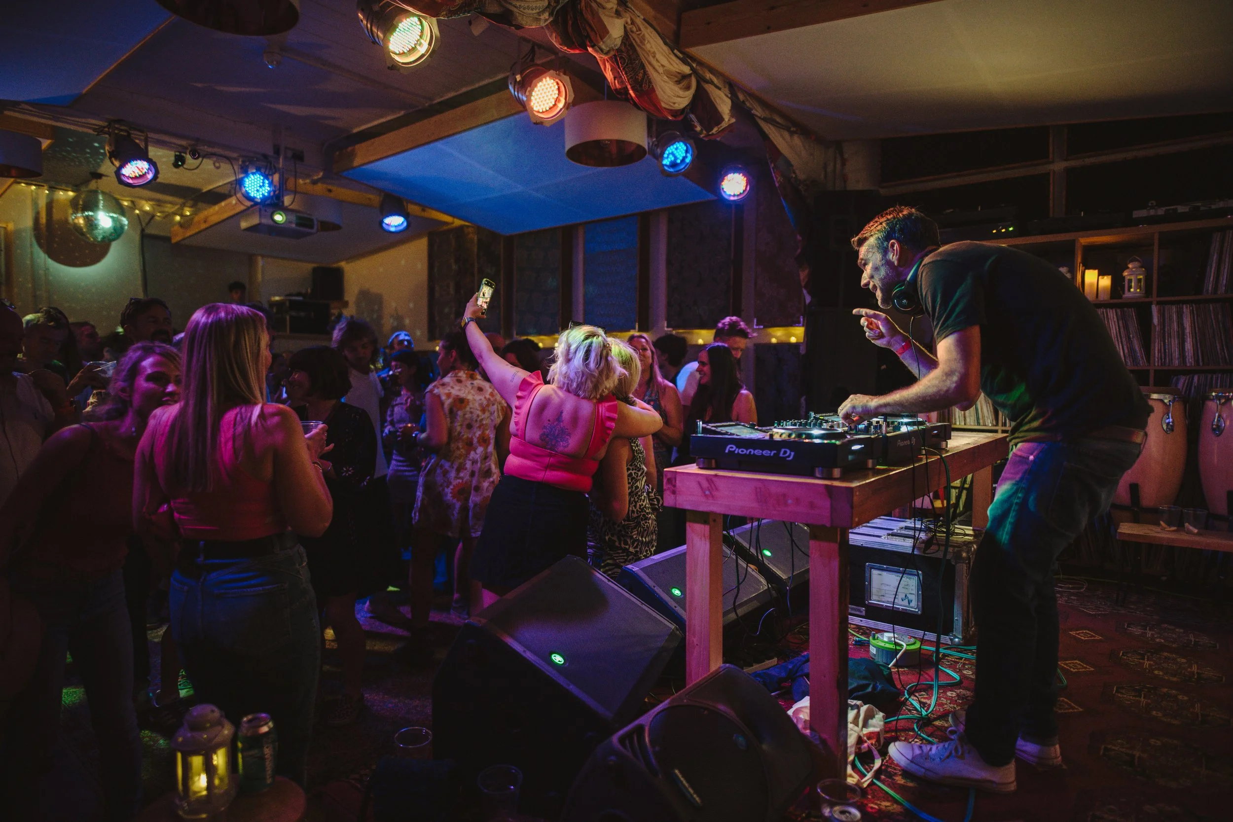 A DJ playing music at a party with people dancing and enjoying themselves in a dimly lit room with colorful lights.