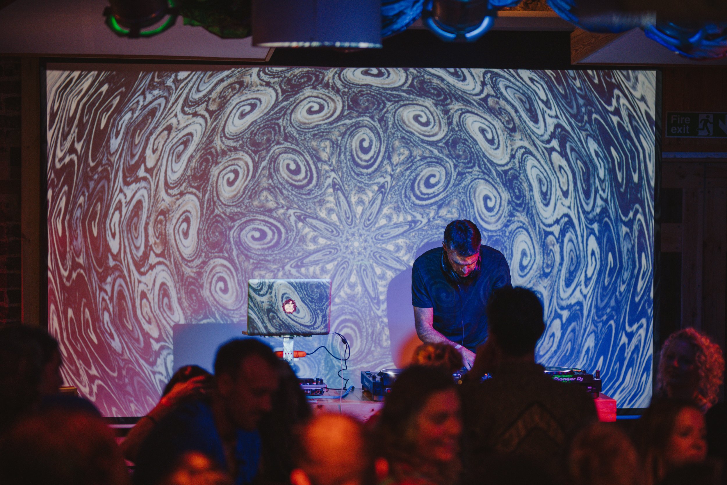 DJ performing in front of a large, patterned projection screen with swirling blue and white designs, and audience members watching.