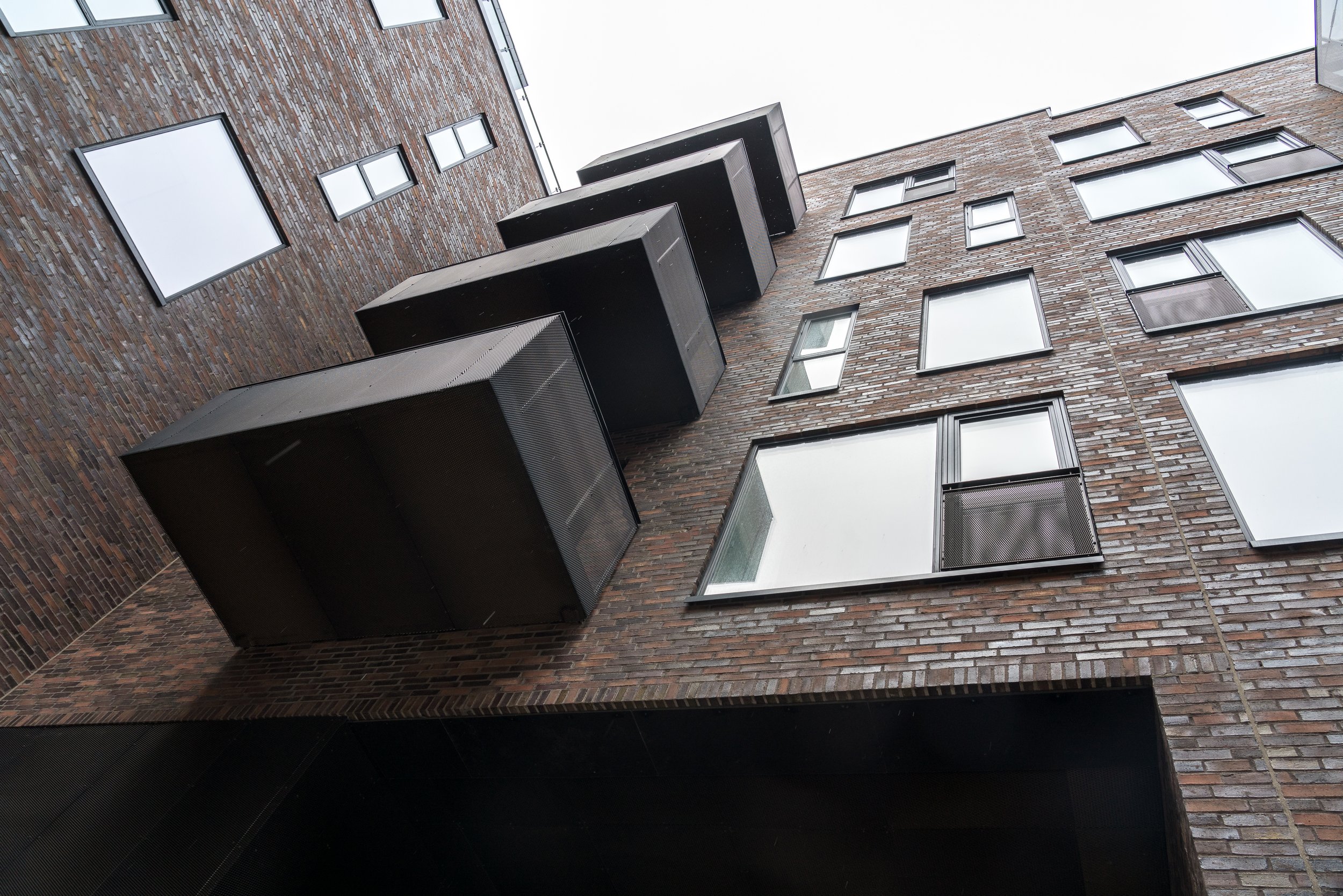 Low-angle view of a modern brick apartment building with black balconies and large windows under a cloudy sky.