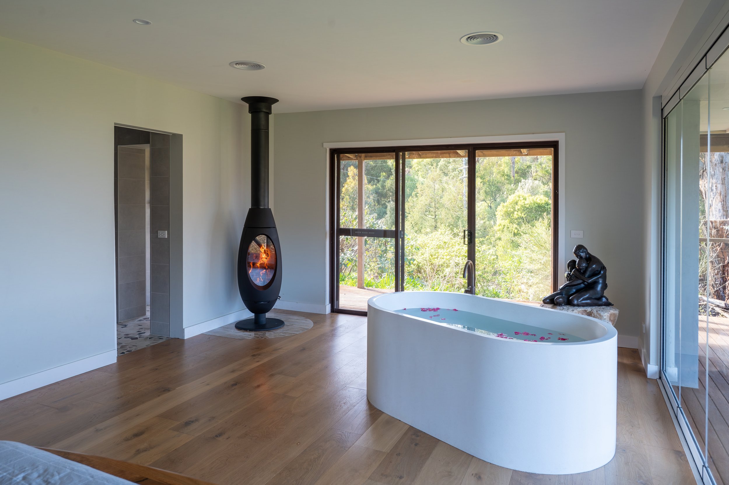 Country Victoria master bedroom featuring a bathtub, bronze sculpture on a hand-selected boulder, a Living Fire fireplace, and wall-to-wall, floor-to-ceiling stacker doors framing valley and distant sea views.