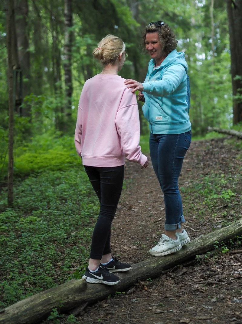 Valmentaja ja NUOTTI-valmennukseen osallistuva nuori seisovat metsässä vastakkain puunrungon päällä. Toisella on vaaleanpunainen paita ja toisella sininen huppari. He katsovat toisiaan.